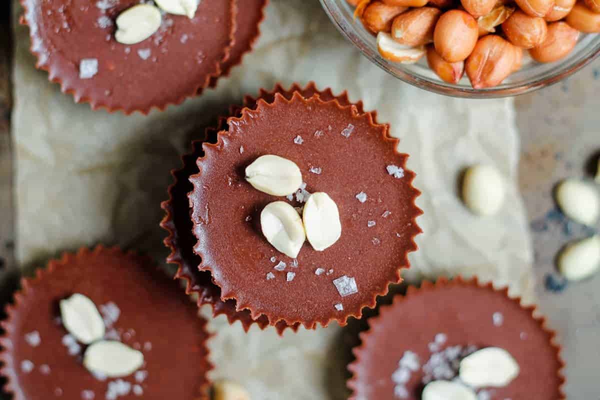 Chocolate peanut butter cups topped with three peanut halves and sea salt flakes, arranged on parchment paper with a bowl of shelled peanuts nearby.