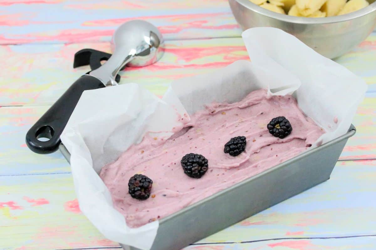 A metal loaf pan lined with parchment paper holds a purple ice cream mixture topped with four blackberries. An ice cream scoop and a metal bowl with sliced bananas are in the background on a pastel-colored surface.