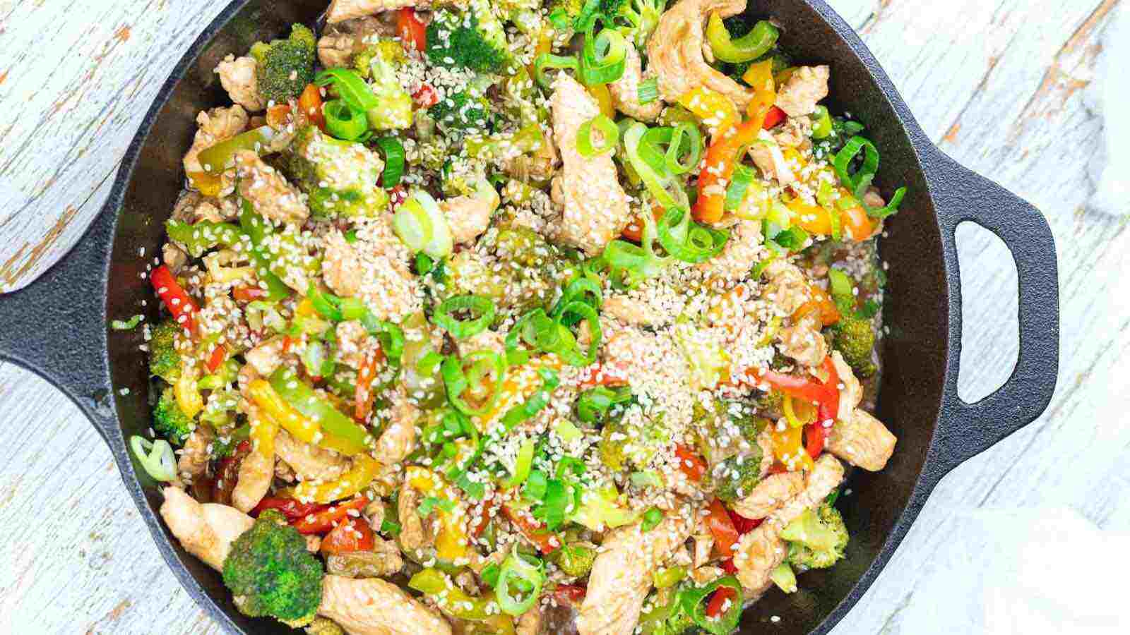A cast iron skillet filled with stir-fried chicken pieces, broccoli, sliced bell peppers, and green onions, topped with sesame seeds. The food is colorful and appears freshly cooked.