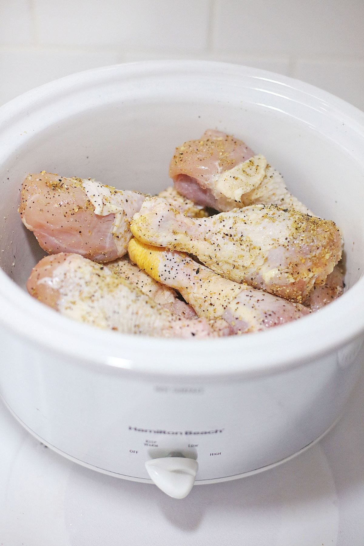 Raw, seasoned chicken drumsticks arranged inside a white slow cooker, ready to be cooked; the chicken is sprinkled with black pepper and spices.