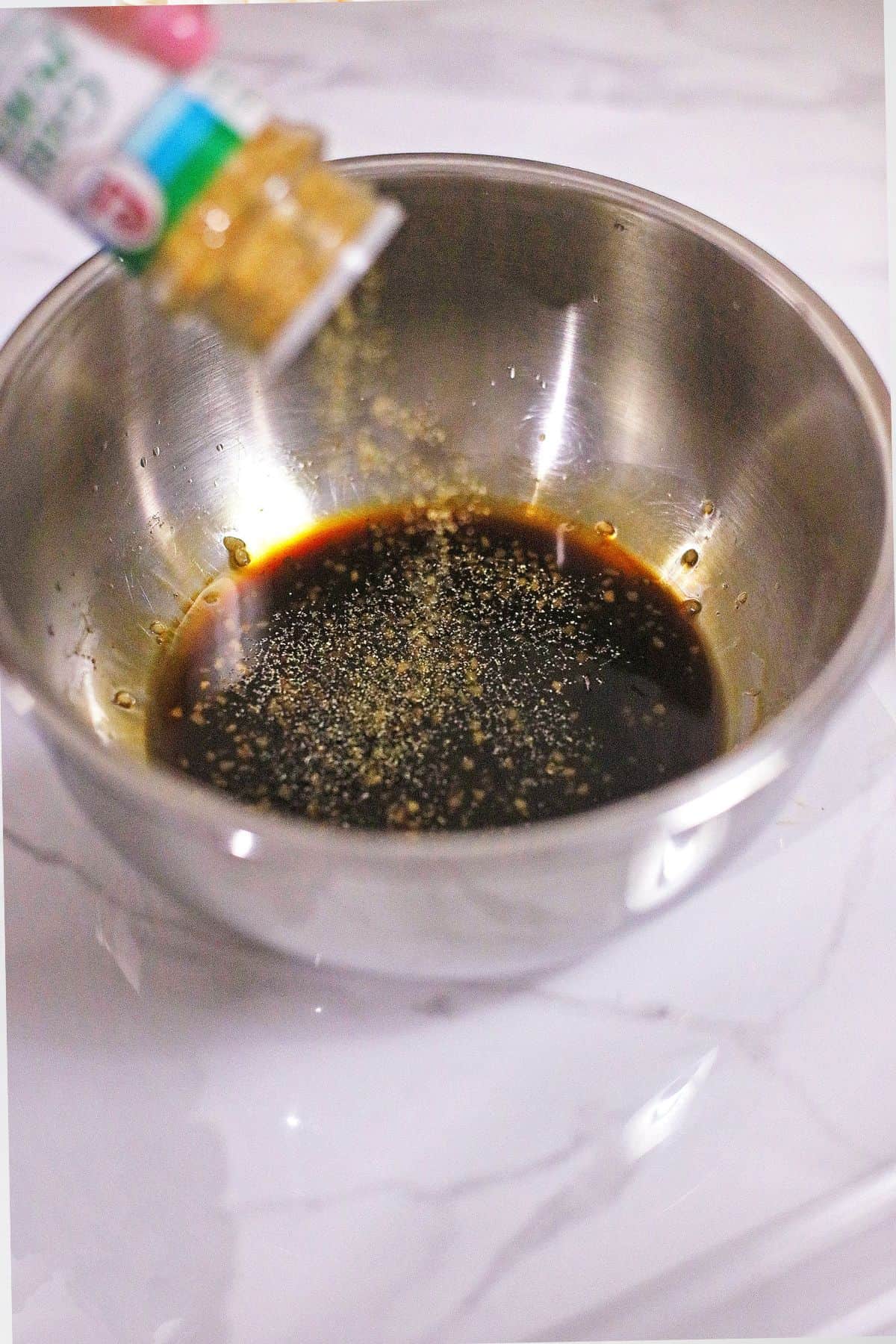 A metal bowl with dark liquid inside is placed on a marble surface. Ground spices are being poured into the bowl from a plastic container above.
