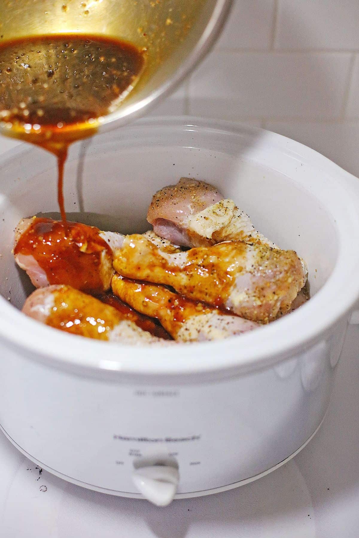 A bowl of sauce is being poured over raw chicken drumsticks seasoned with spices in a white slow cooker on a kitchen counter.