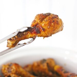 A pair of metal tongs holding a seasoned and cooked chicken drumstick above a bowl containing more chicken drumsticks.