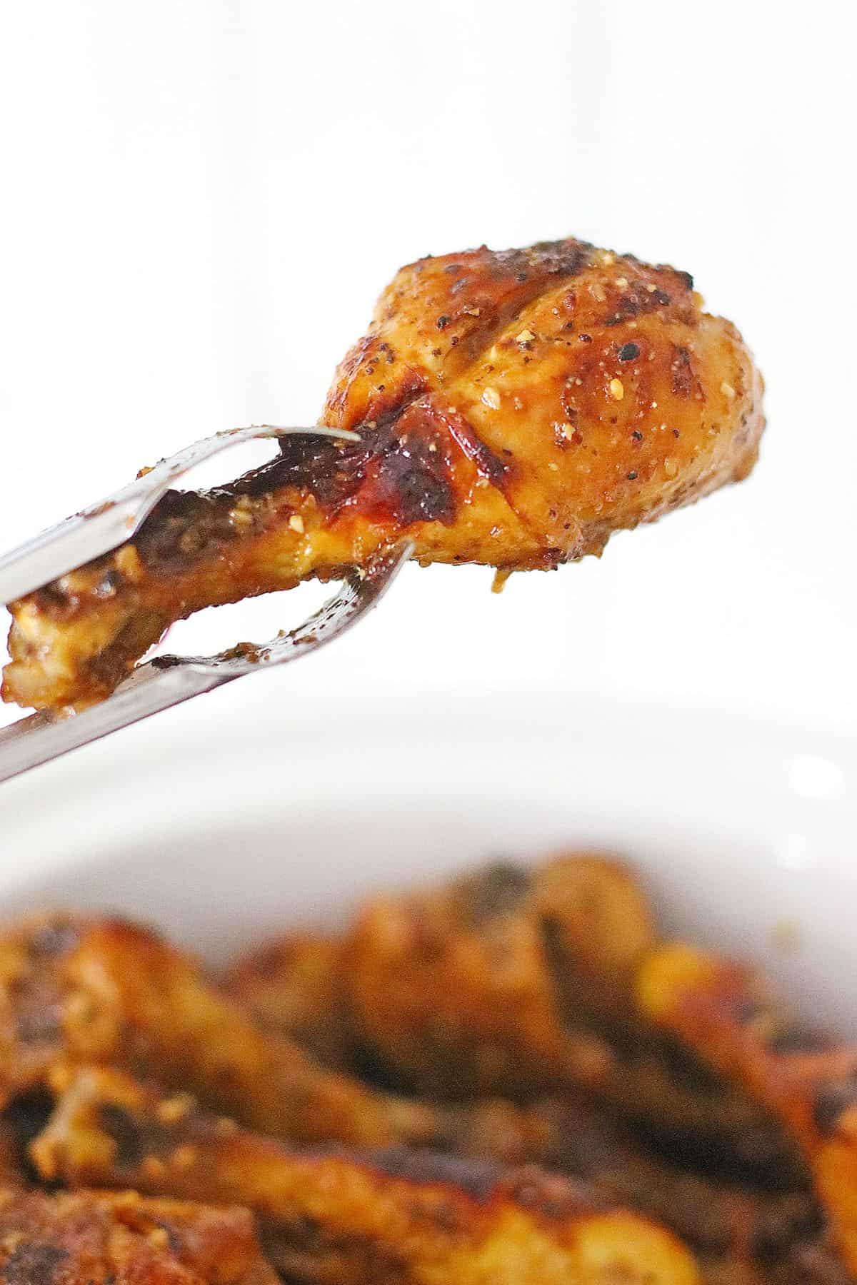 A metal tong holds a cooked, seasoned chicken drumstick above a bowl filled with more drumsticks, against a plain white background.