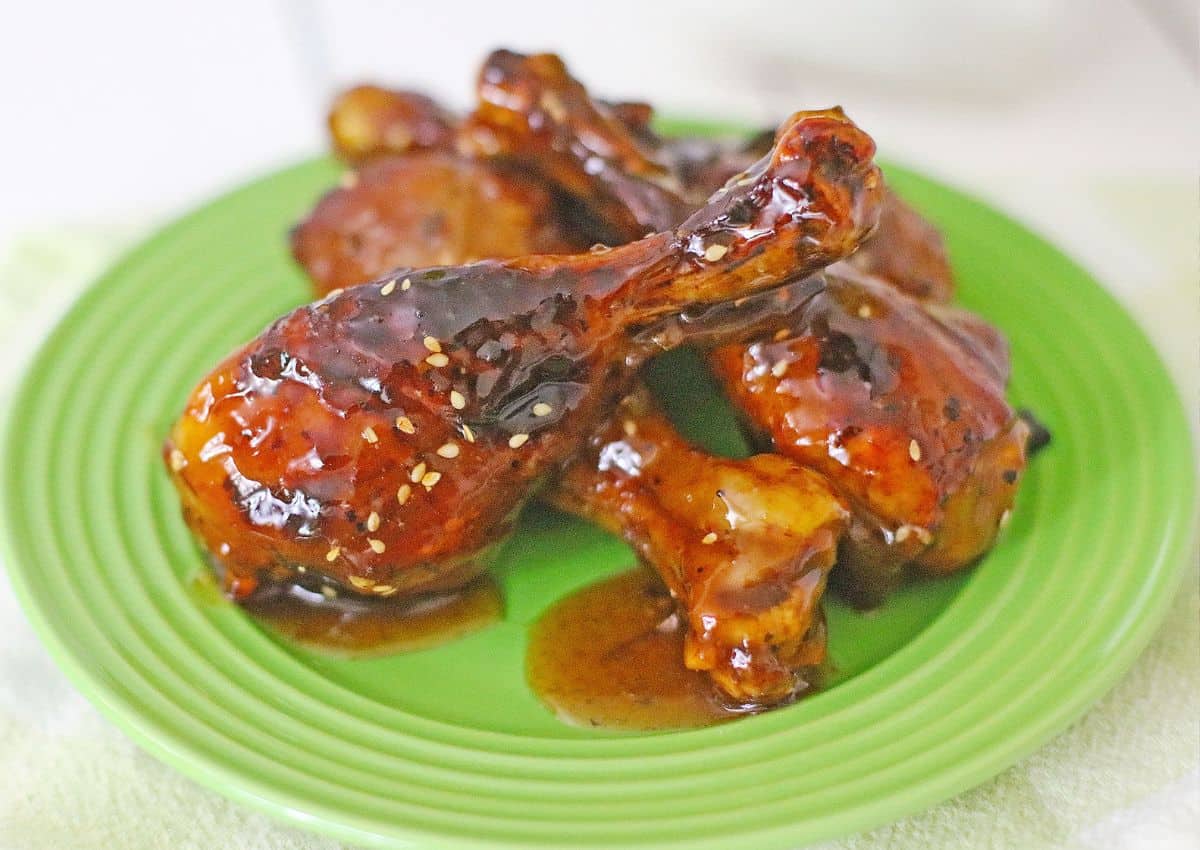 Four glazed chicken drumsticks coated in a dark, shiny sauce with sesame seeds are arranged on a green plate, with some sauce pooled underneath them.