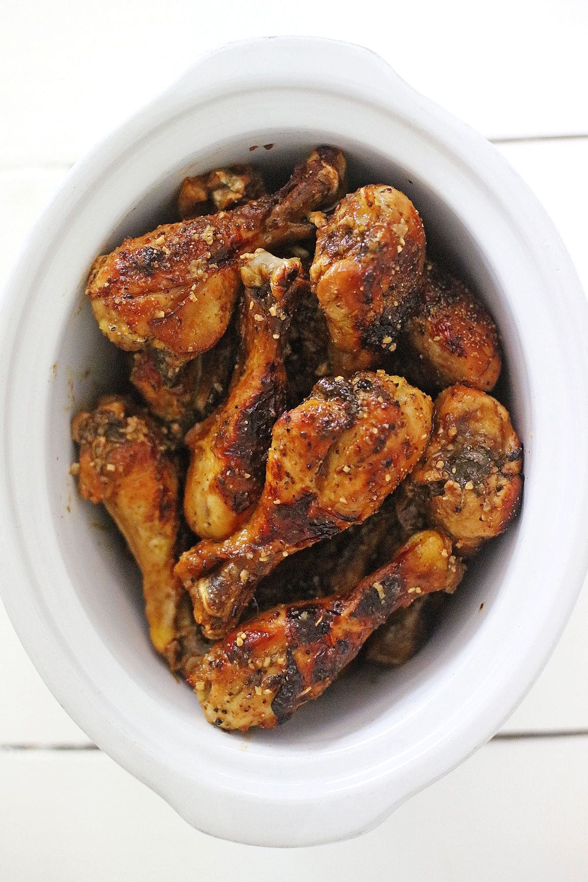 A white slow cooker filled with seasoned and cooked chicken drumsticks, viewed from above on a white surface.