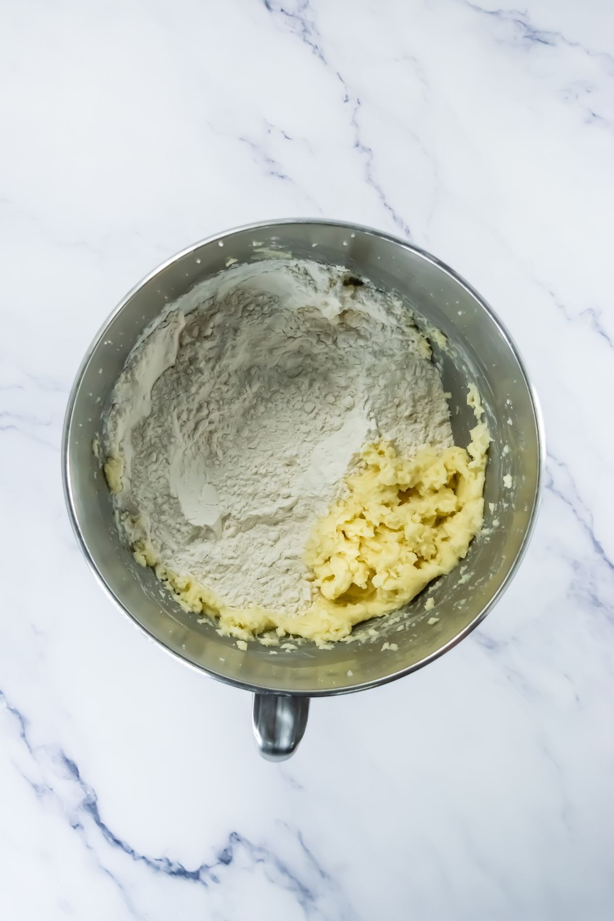 A metal mixing bowl on a marble surface containing partially mixed dough, with white flour on one side and creamed butter and sugar on the other.