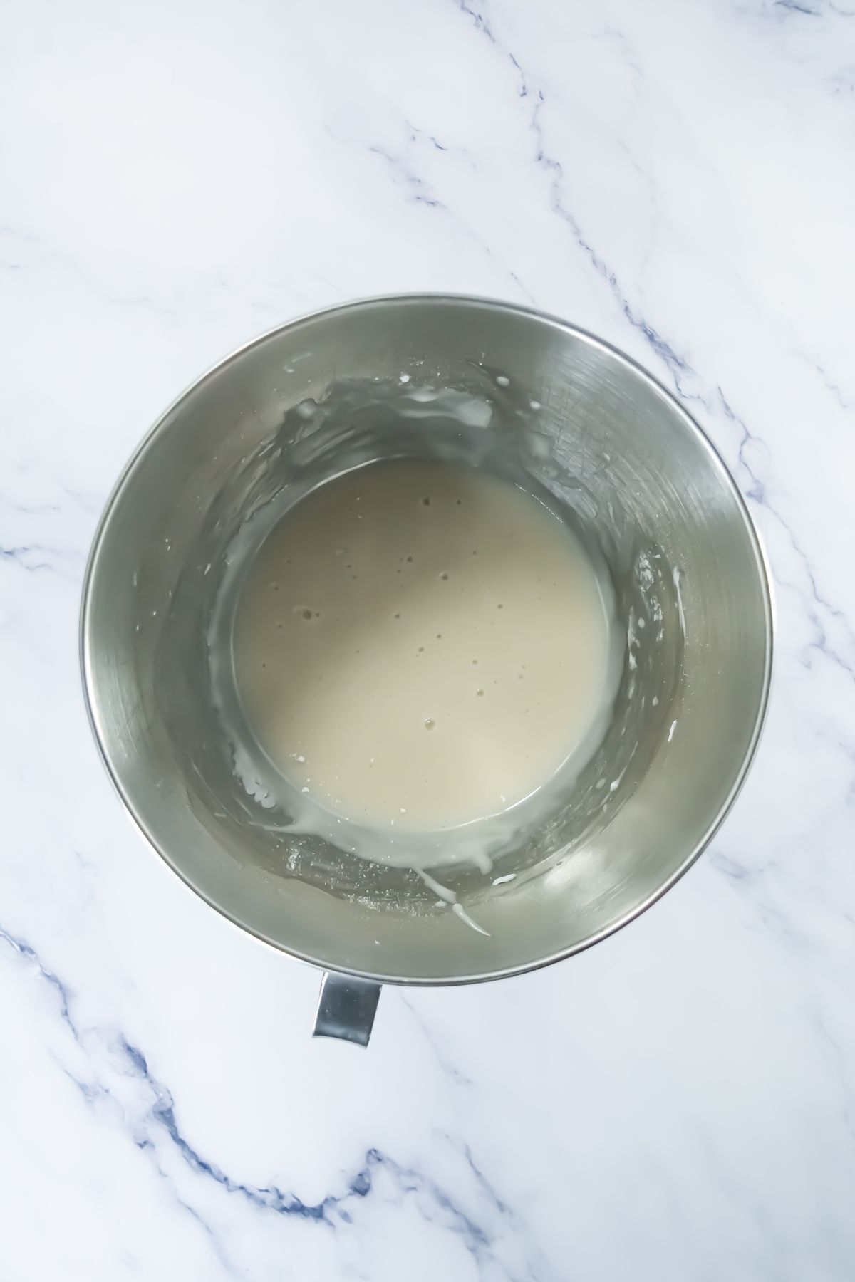 A stainless steel mixing bowl containing a light beige batter sits on a white marble surface. Some batter is splattered along the inner sides of the bowl.