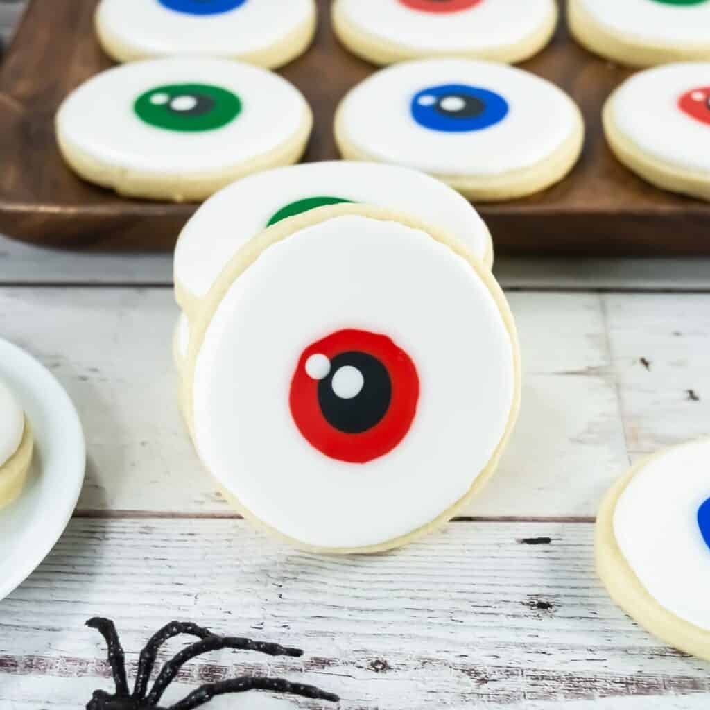 A round sugar cookie with white icing and a red and black circle in the center, resembling an eyeball. Other cookies with similar designs in different colors are in the background on a tray.