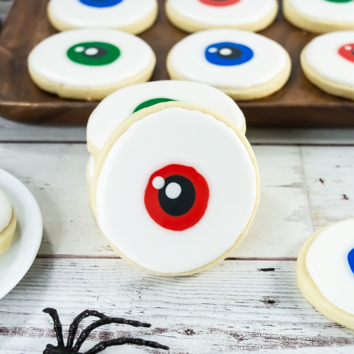 A round sugar cookie with white icing and a red and black circle in the center, resembling an eyeball. Other cookies with similar designs in different colors are in the background on a tray.