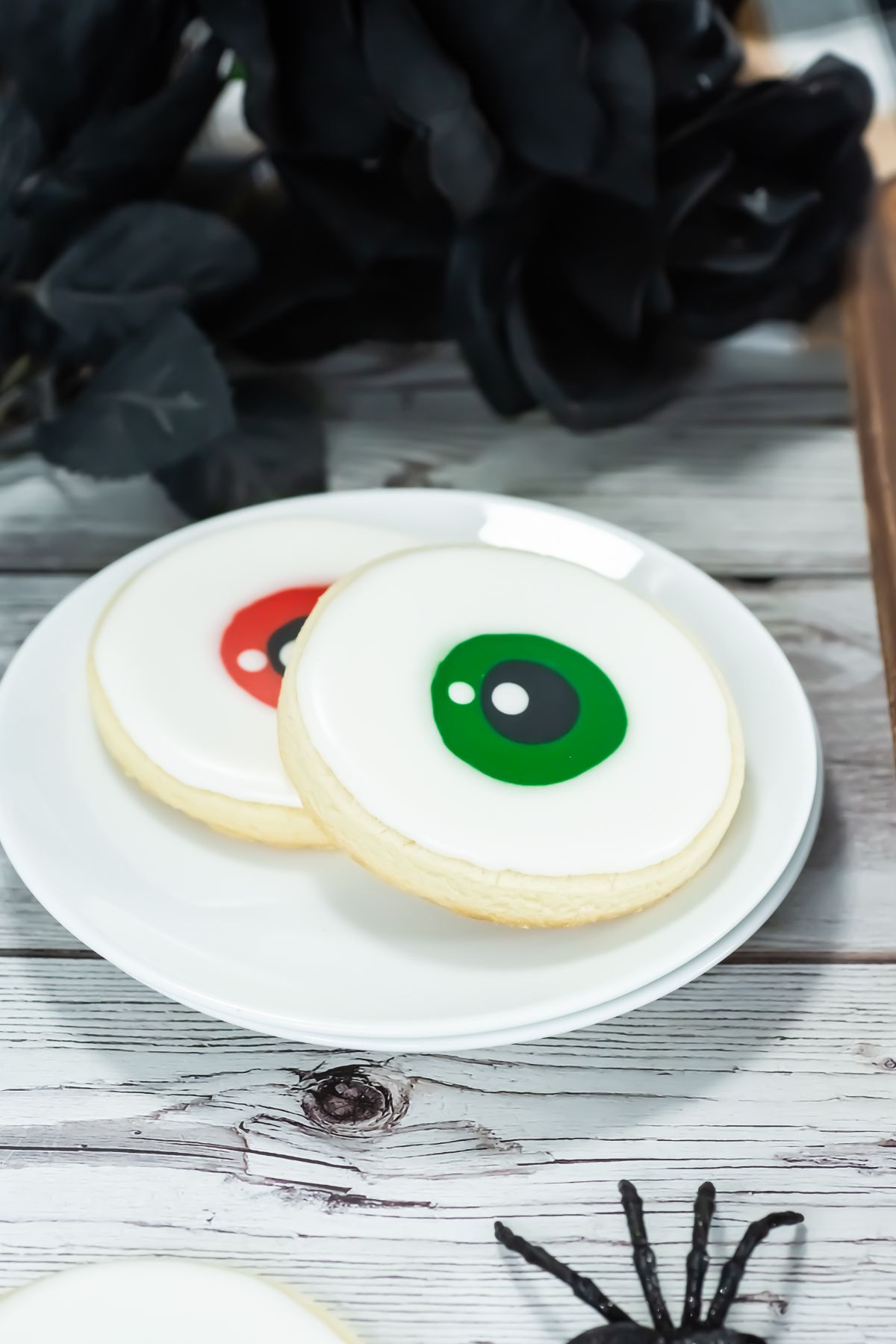 Two cookies decorated with white icing and colored circles resembling eyeballs sit on a white plate. The background includes dark artificial leaves and part of a plastic spider on a wooden surface.