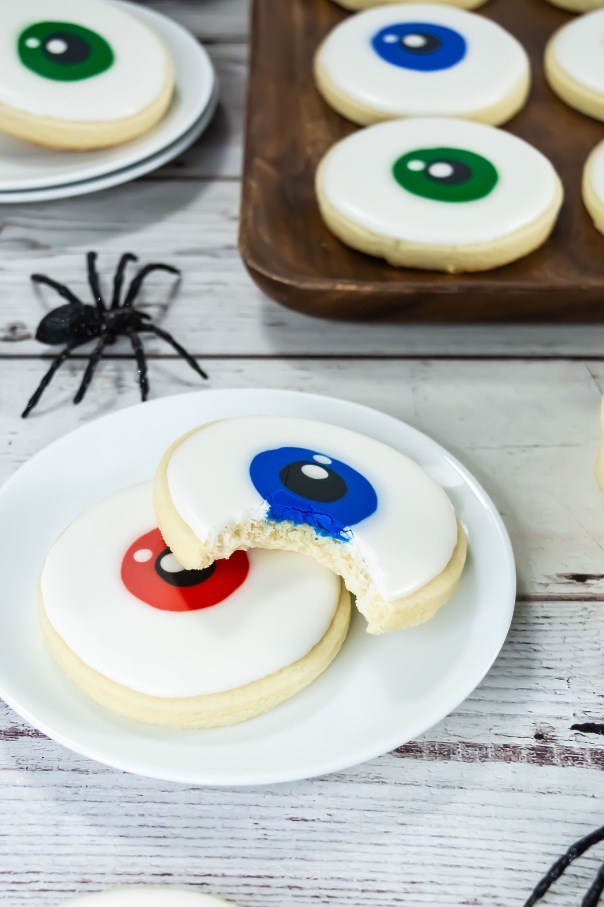 A white plate holds two round cookies decorated to look like eyeballs, one with a red iris and one with a blue iris that has a bite taken out of it. Other similar cookies and a toy black spider are in the background.
