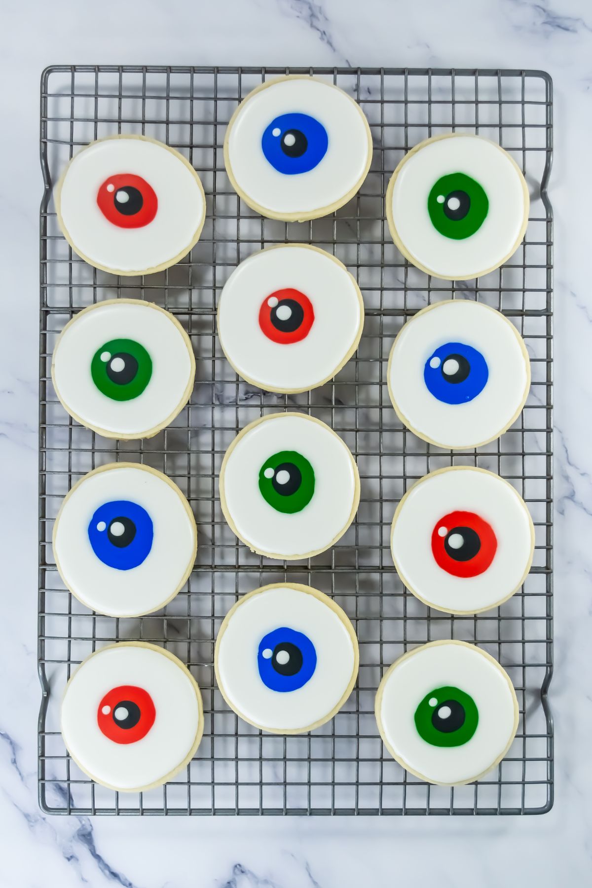 Twelve round cookies with white icing and colored circles resembling eyeballs—red, green, and blue—arranged in rows on a wire cooling rack set on a white marble surface.