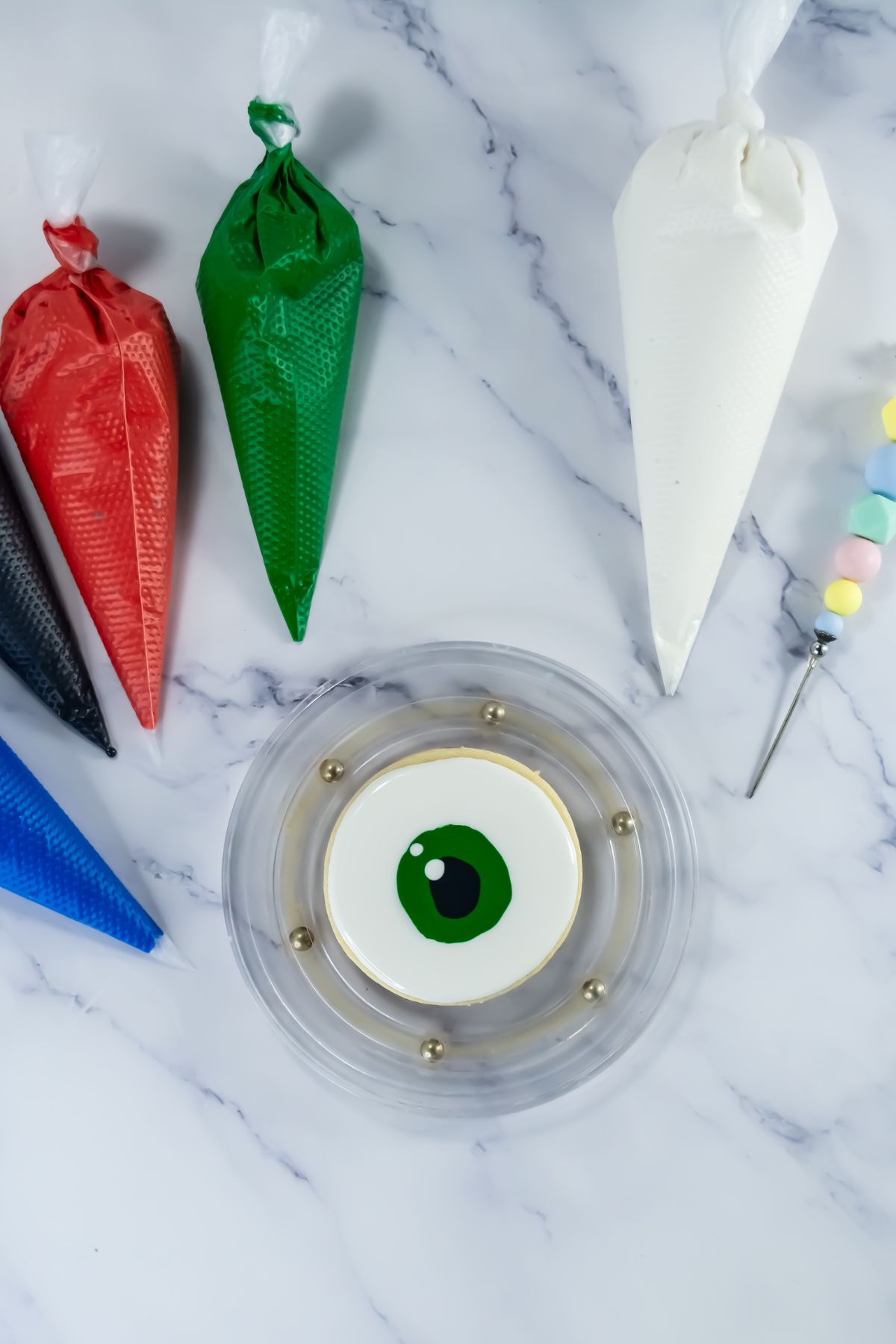 A round cookie decorated with a green and black eye design sits on a clear plate, surrounded by piping bags with red, green, black, blue, and white icing, and a decorating tool with pastel candy beads.