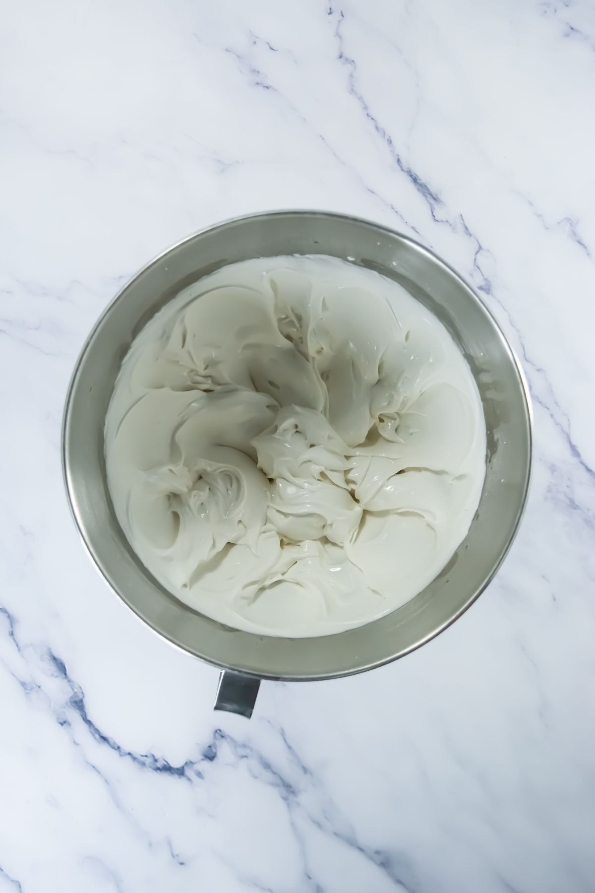 A stainless steel mixing bowl filled with smooth, whipped white frosting sits on a white marble surface with gray veining.