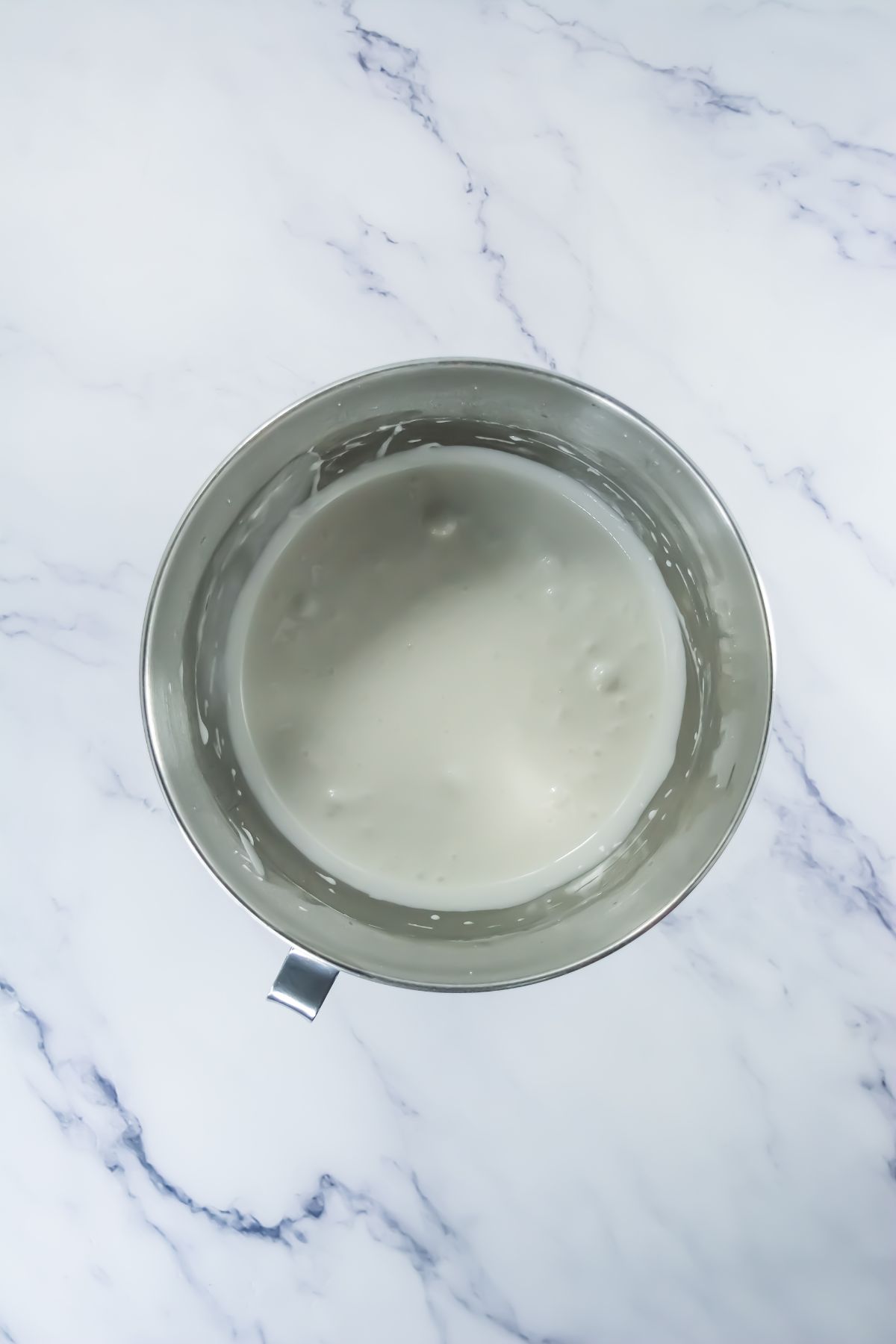 A metal mixing bowl containing a smooth, white batter or mixture sits on a white marble surface.