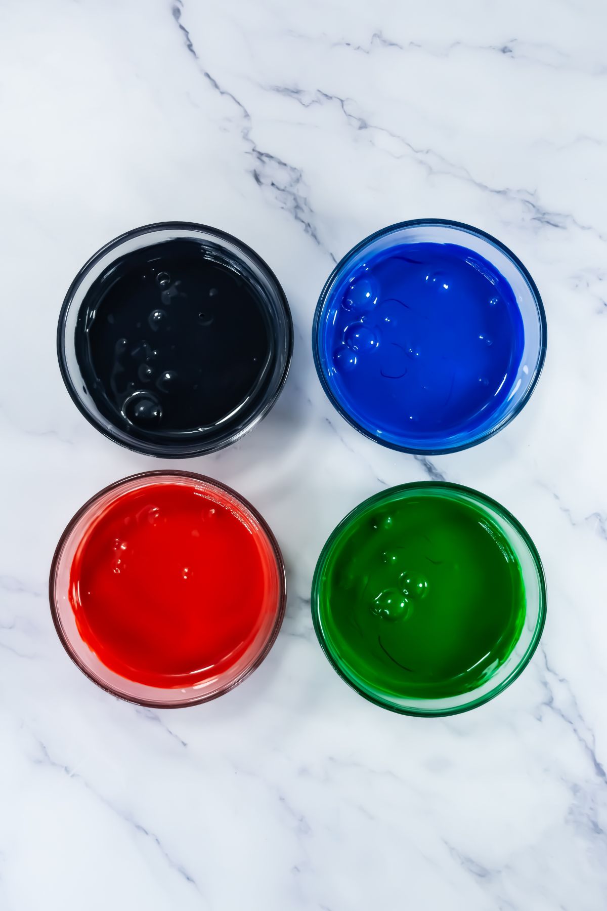 Four glass bowls filled with black, blue, red, and green liquid food coloring are arranged in a square on a white marble surface.