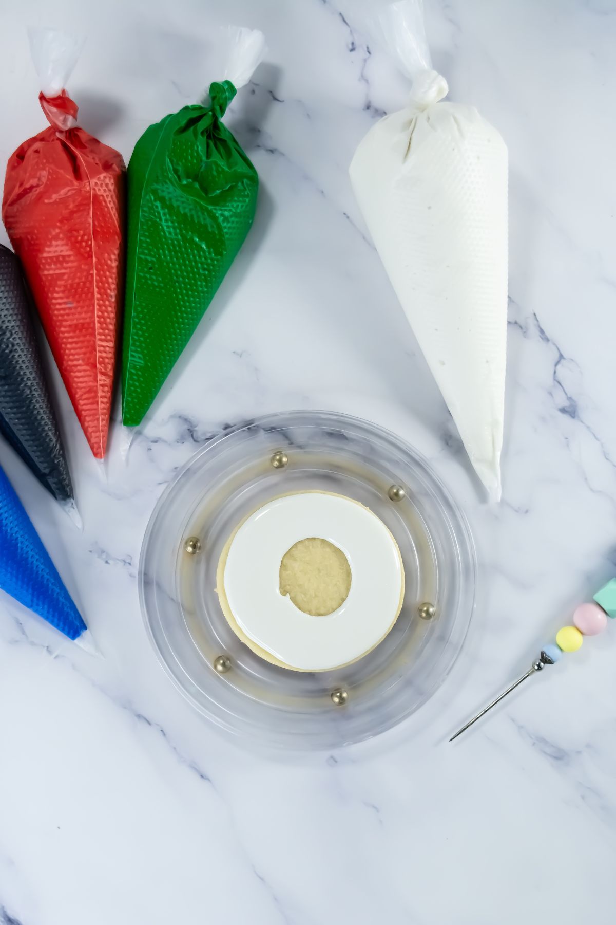 A single round cookie with a white icing border sits on a clear tray. Around it are piping bags filled with red, green, white, black, and blue icing, and a tool with a decorative handle.