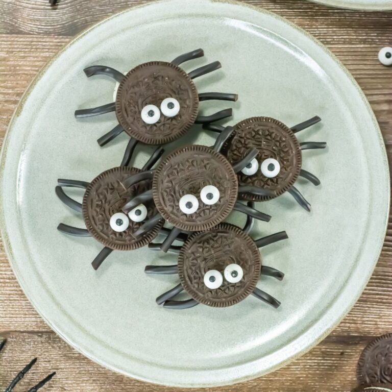 A plate with five cookie sandwiches decorated to look like spiders, each with candy eyes and black licorice pieces for legs, placed on a wooden surface.