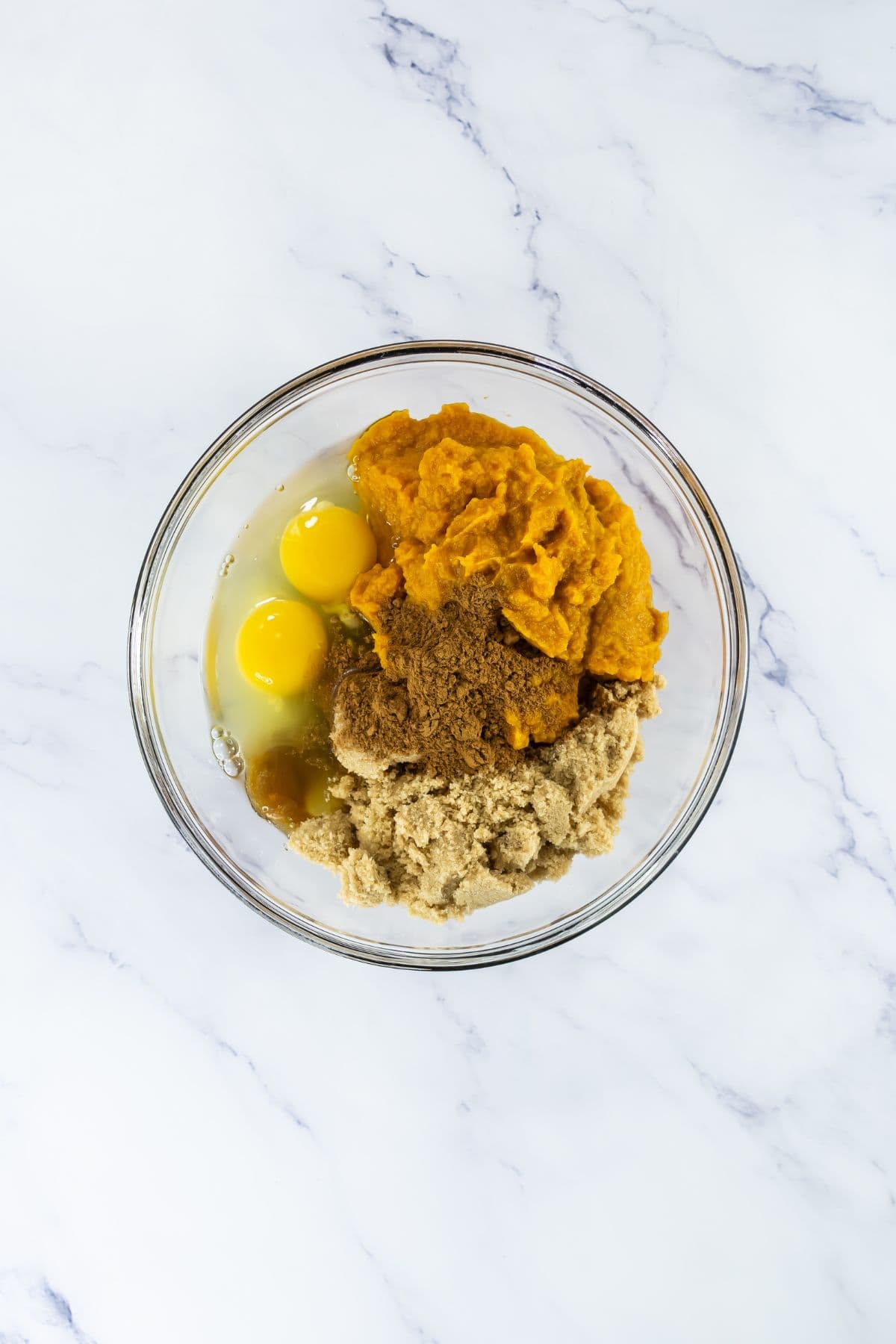A glass mixing bowl containing two eggs, pumpkin puree, brown sugar, and granulated sugar on a white marble surface.