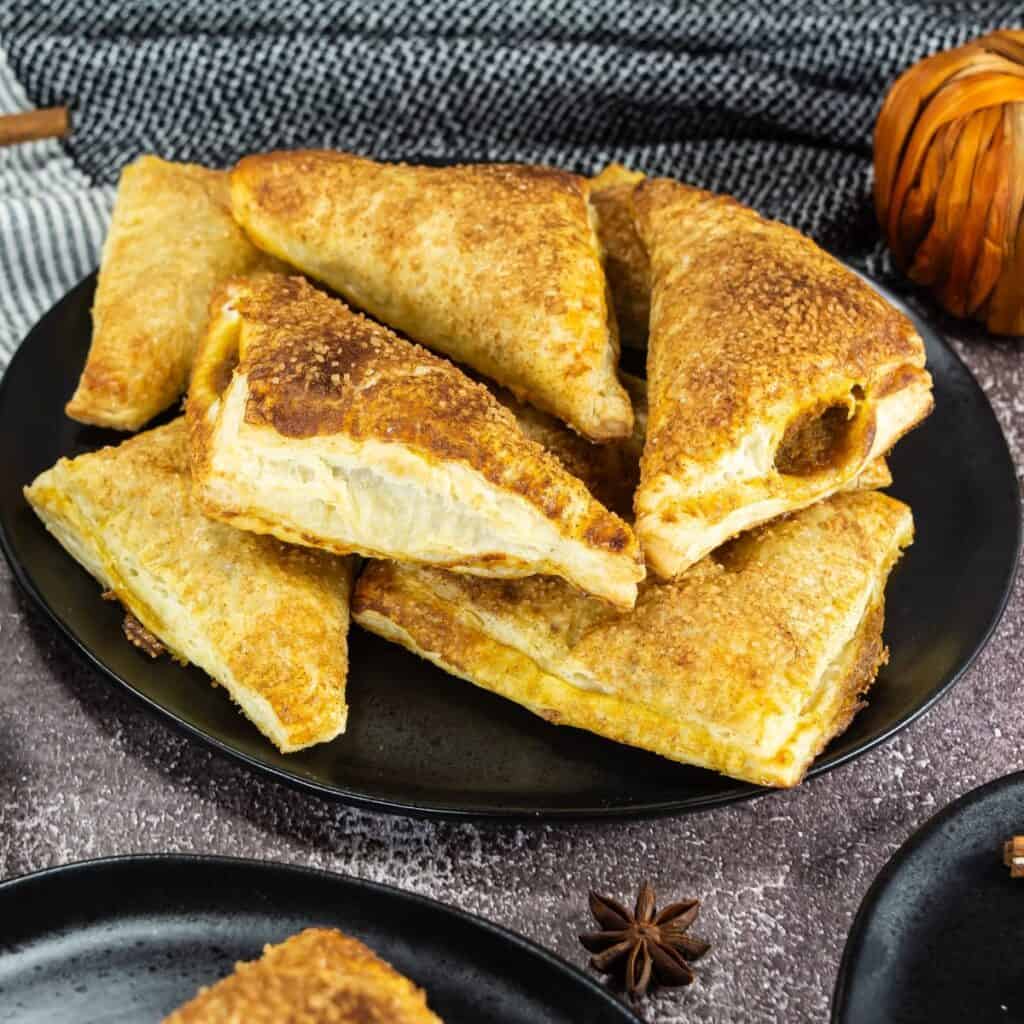 A black plate holds several golden-brown, triangular puff pastry turnovers. The pastries are stacked and arranged neatly. A textured gray cloth and decorative items are in the background.