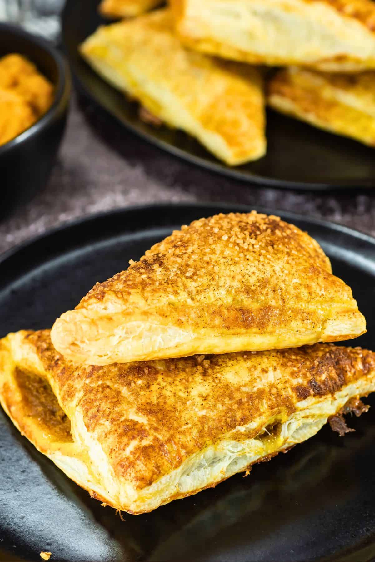 Two golden-brown puff pastry turnovers with a slightly flaky texture are stacked on a black plate. More pastries are visible in the background along with a dark bowl.