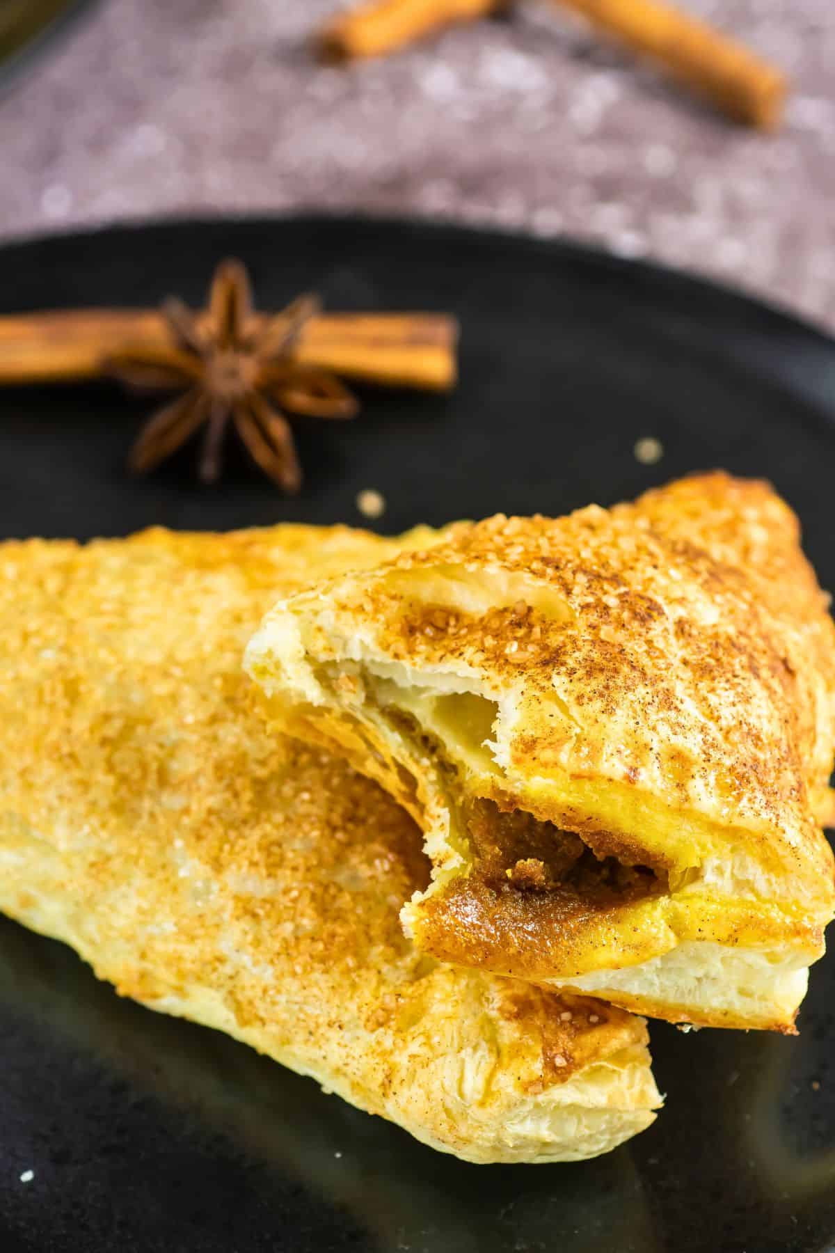 A close-up of two golden-brown puff pastry turnovers on a black plate, one partially broken open to show a spiced apple filling. In the background, whole cinnamon sticks and star anise are visible.