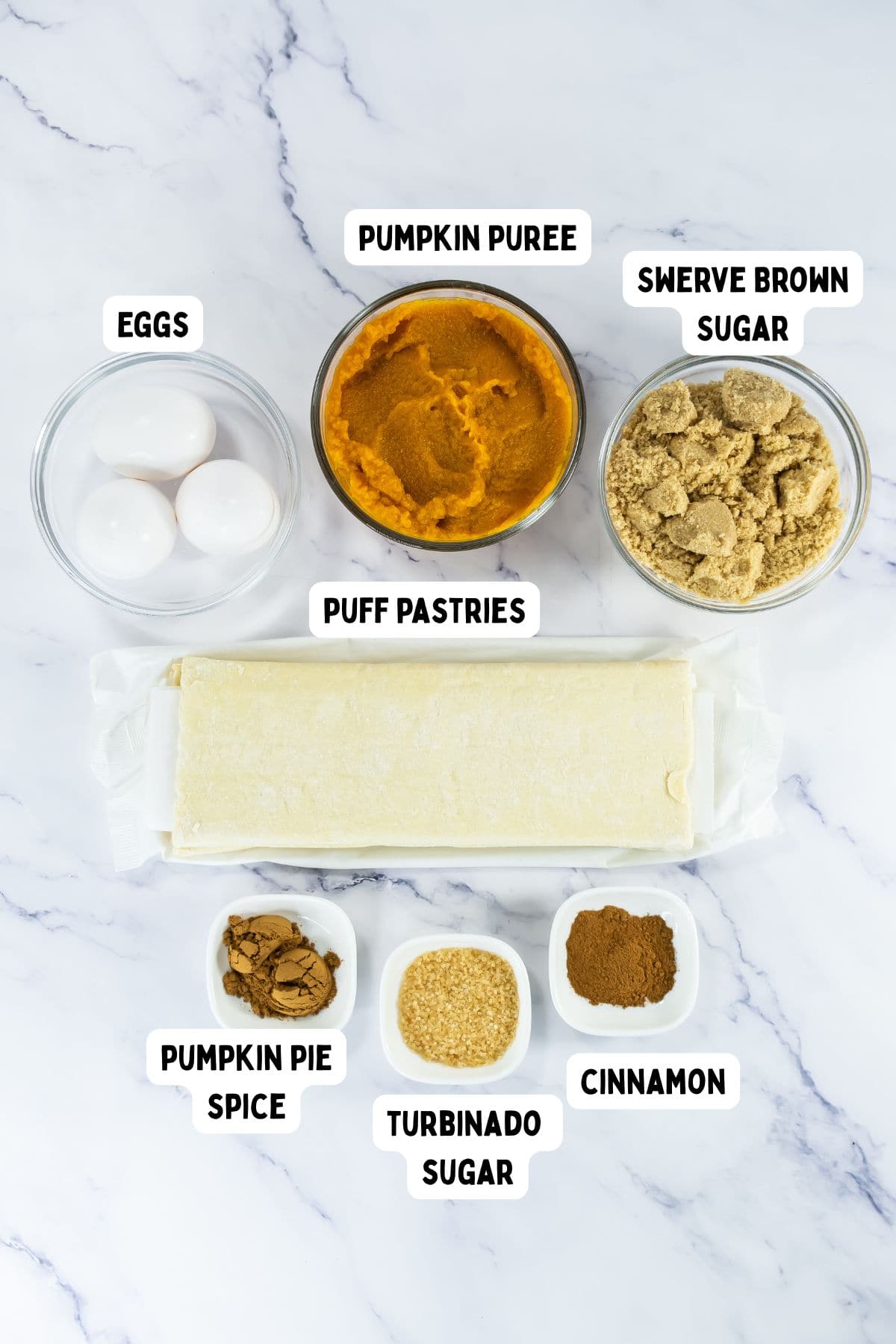 A marble surface displays bowls of eggs, pumpkin puree, Swerve brown sugar, pumpkin pie spice, turbinado sugar, cinnamon, and a sheet of puff pastry, each ingredient clearly labeled.