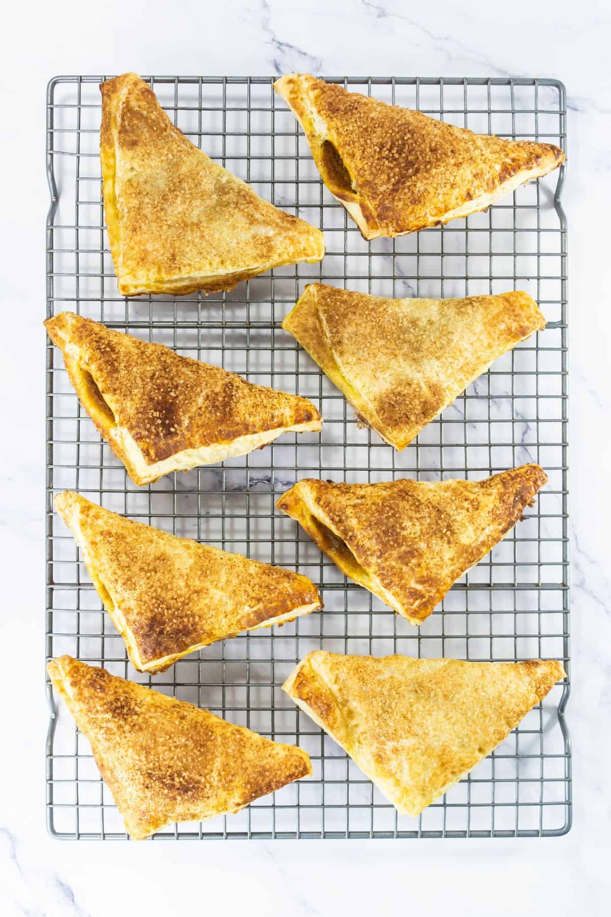 Nine golden-brown, triangular pastries are arranged on a wire cooling rack over a white marble surface. The pastries have a crisp, flaky texture and are evenly spaced on the rack.