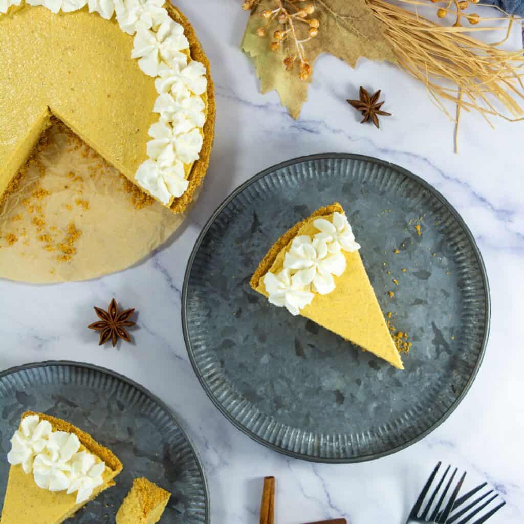 A slice of pumpkin pie with whipped cream sits on a gray plate. The rest of the pie, with a slice missing, is on a separate plate. Decorative autumn leaves and spices are scattered on a white marble surface.