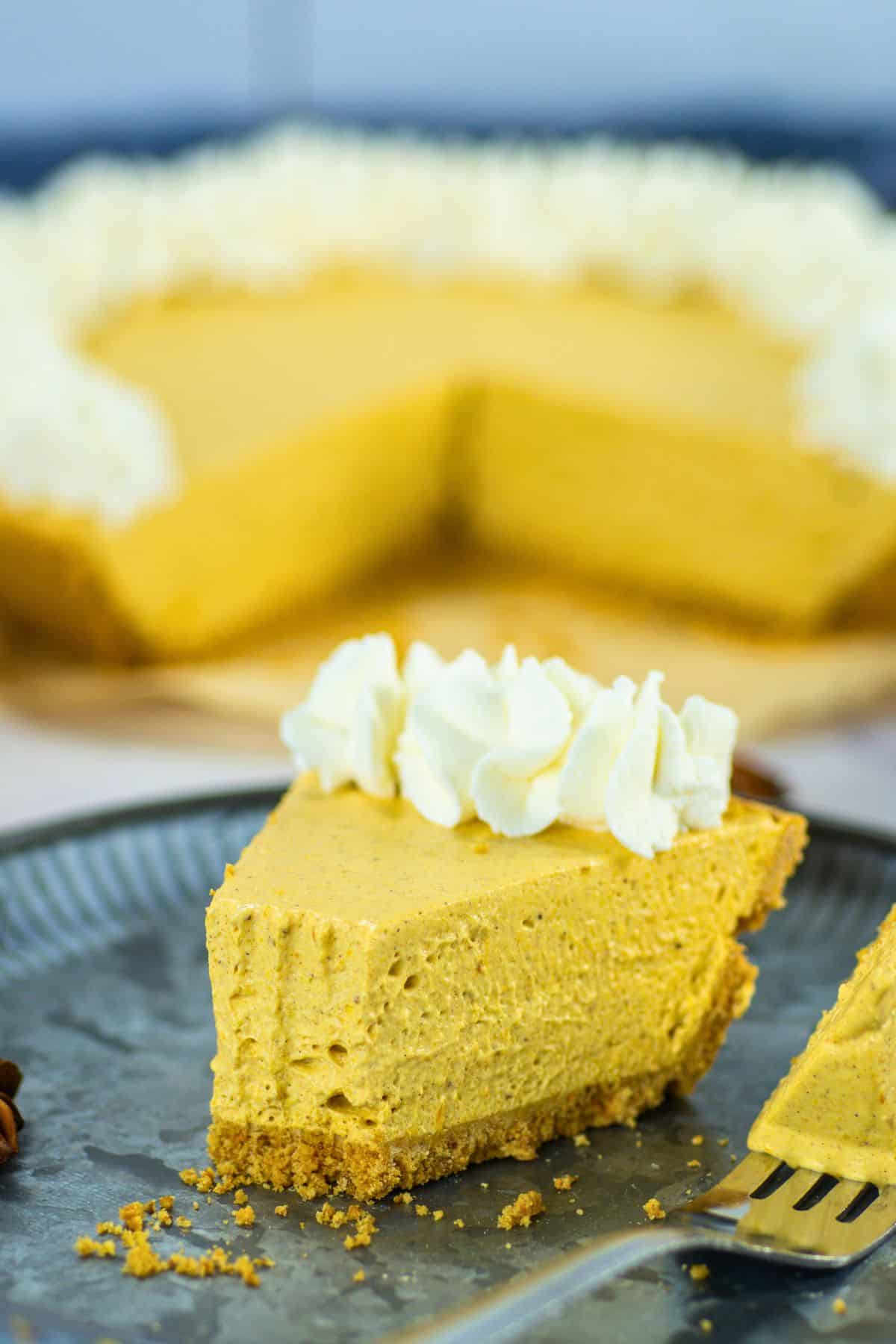 A slice of pumpkin cheesecake with a graham cracker crust and whipped cream topping sits on a dark plate with a fork, reminiscent of a No Bake Pumpkin Pie. The rest of the cheesecake is in the background with a slice missing.