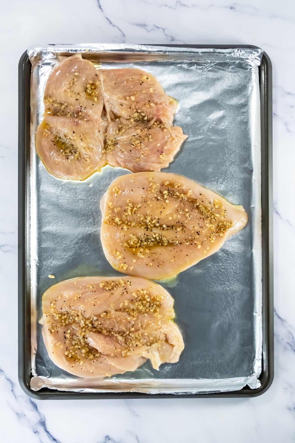 Three raw, marinated chicken breasts with visible herbs and spices are placed on a foil-lined baking sheet, ready for cooking. The baking sheet sits on a white marble surface.
