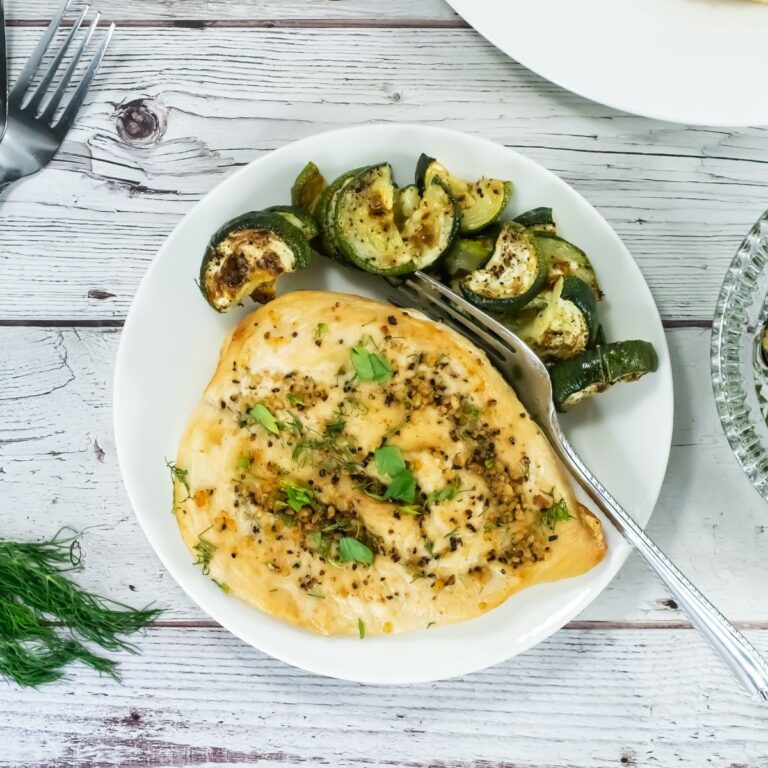 A white plate with a cooked, seasoned chicken breast garnished with herbs and a side of roasted zucchini slices, placed on a rustic white wooden table with a fork and some fresh dill nearby.