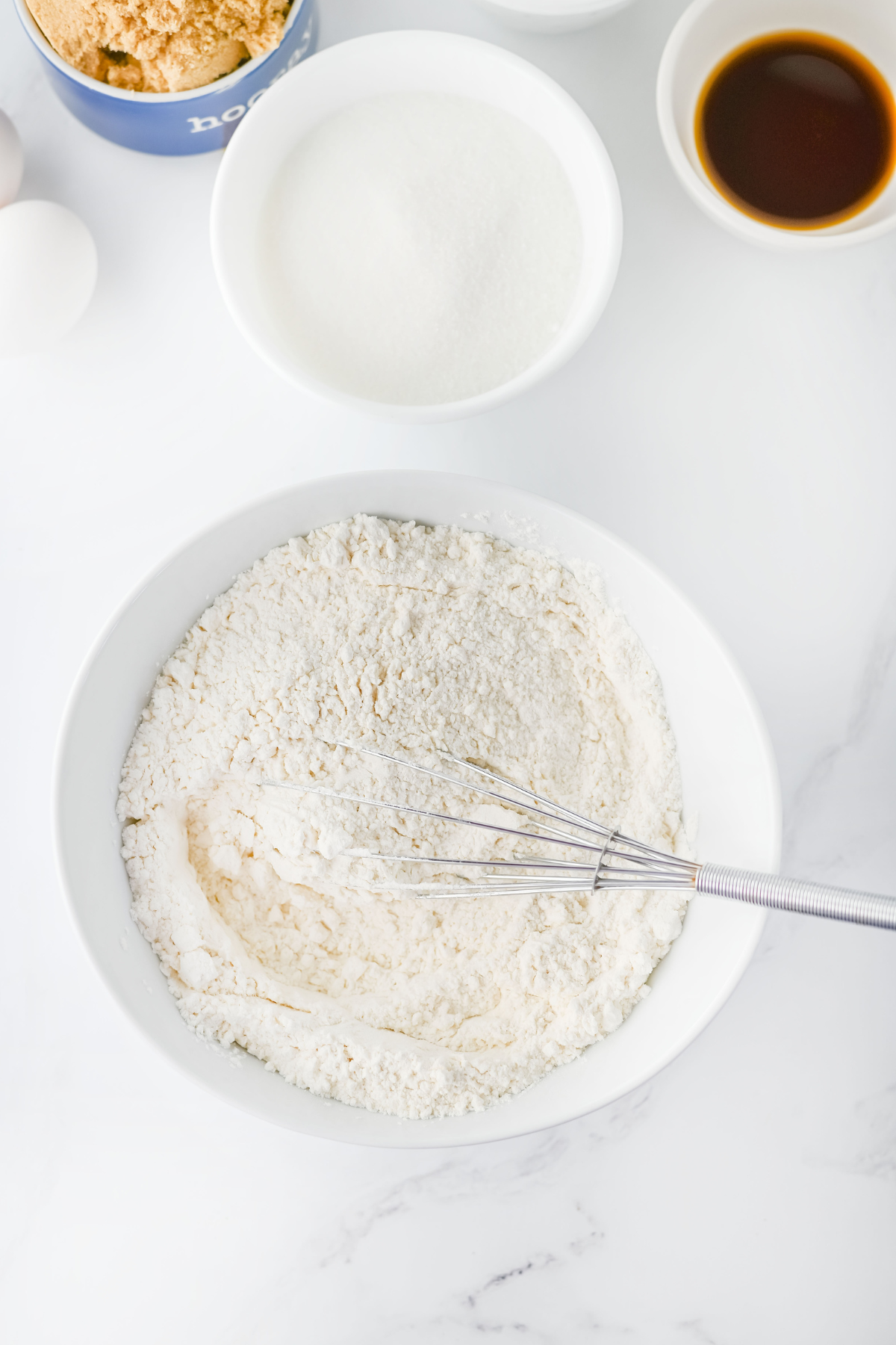 A metal whisk rests in a bowl of flour mixture. Surrounding the bowl are a dish of granulated sugar, a bowl of brown sugar, a small bowl of vanilla extract, and two eggs on a white surface.