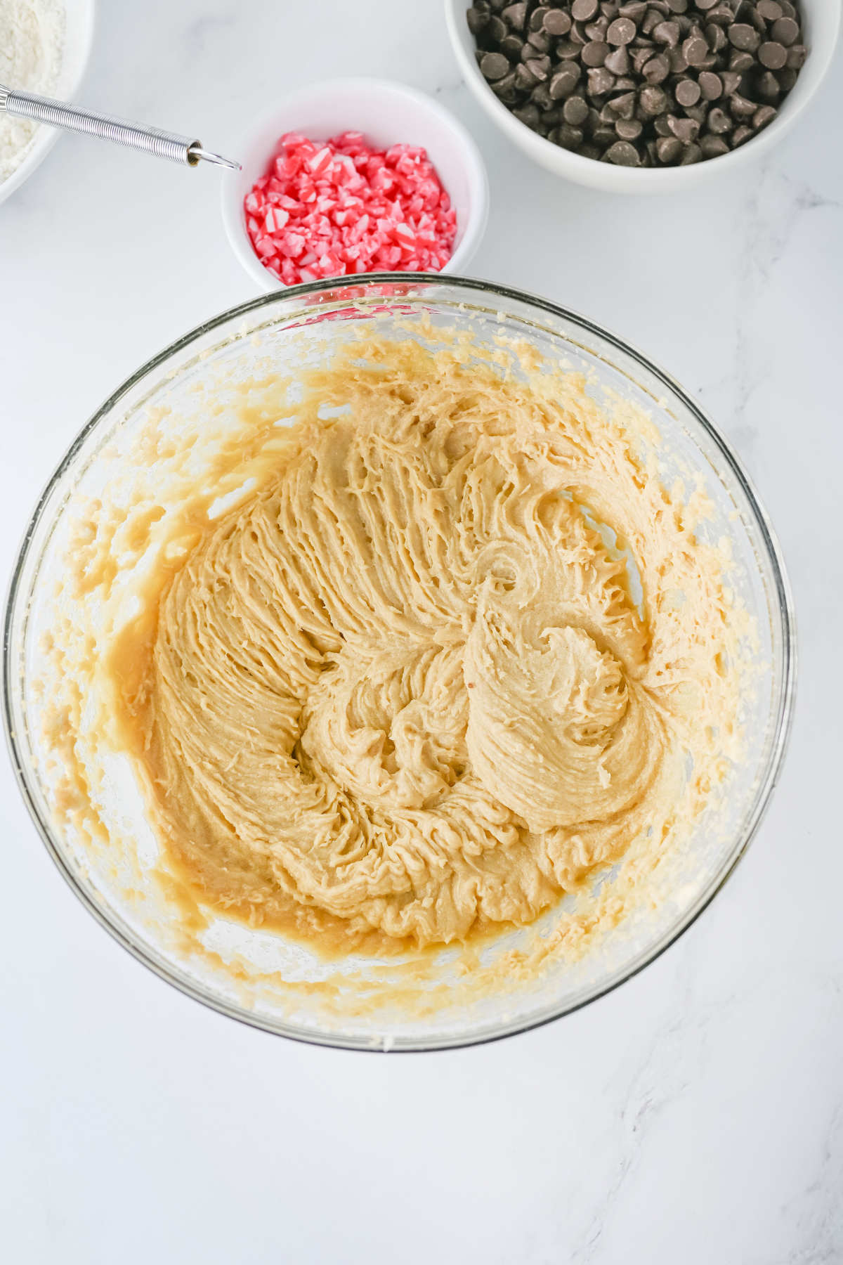 A glass bowl filled with creamed cookie dough sits on a white surface. Nearby are a bowl of chocolate chips and a bowl of crushed red candy pieces. A metal spoon is partially visible in the dough.