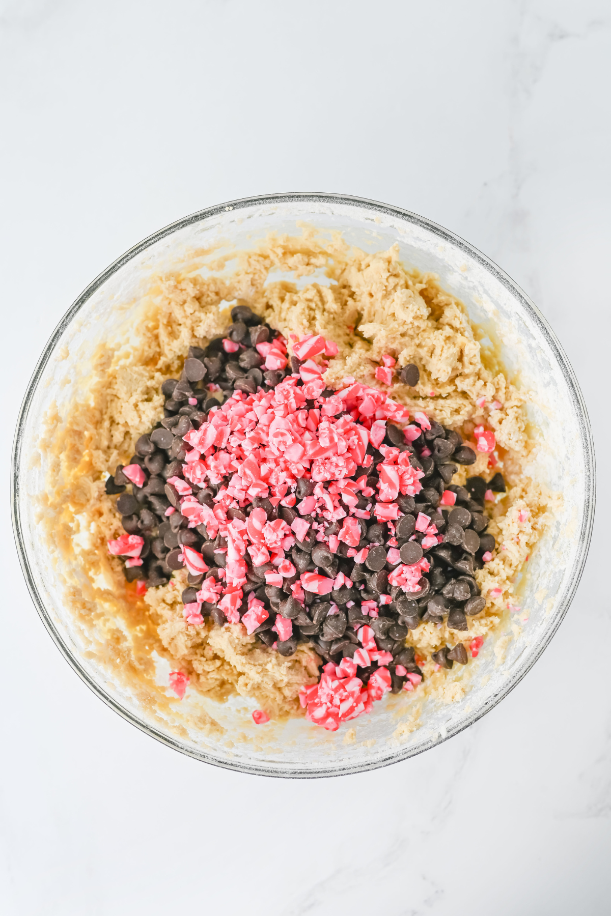 A glass bowl filled with cookie dough, topped with a pile of chocolate chips and a pile of crushed pink candy pieces, on a white surface.