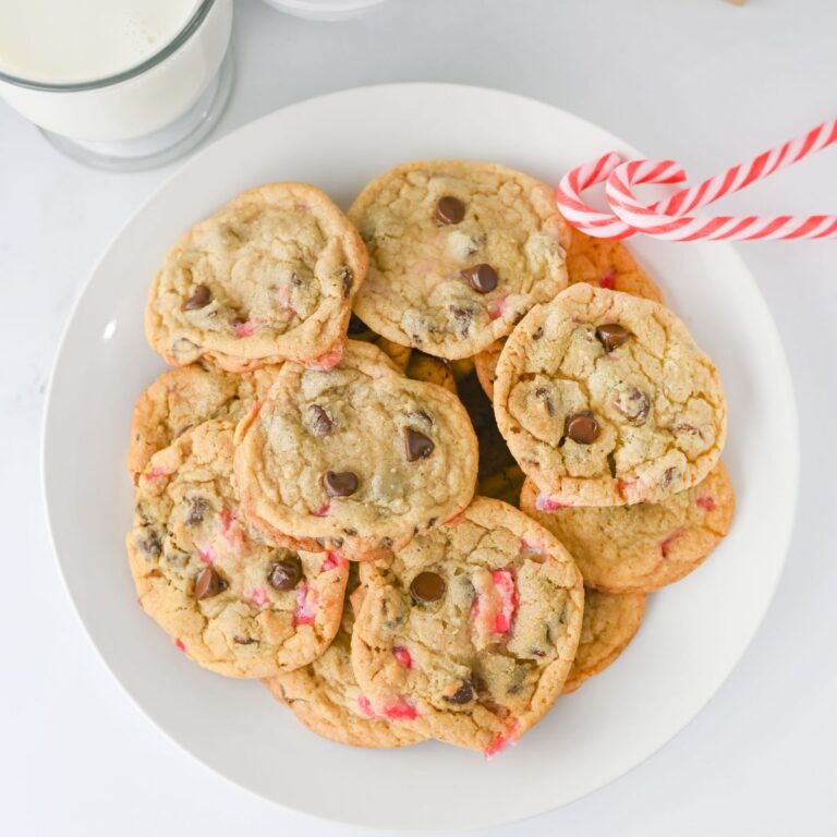 A white plate with Peppermint Chocolate Chip Cookies and pieces of peppermint, next to two red and white candy canes, with a glass of milk in the background.