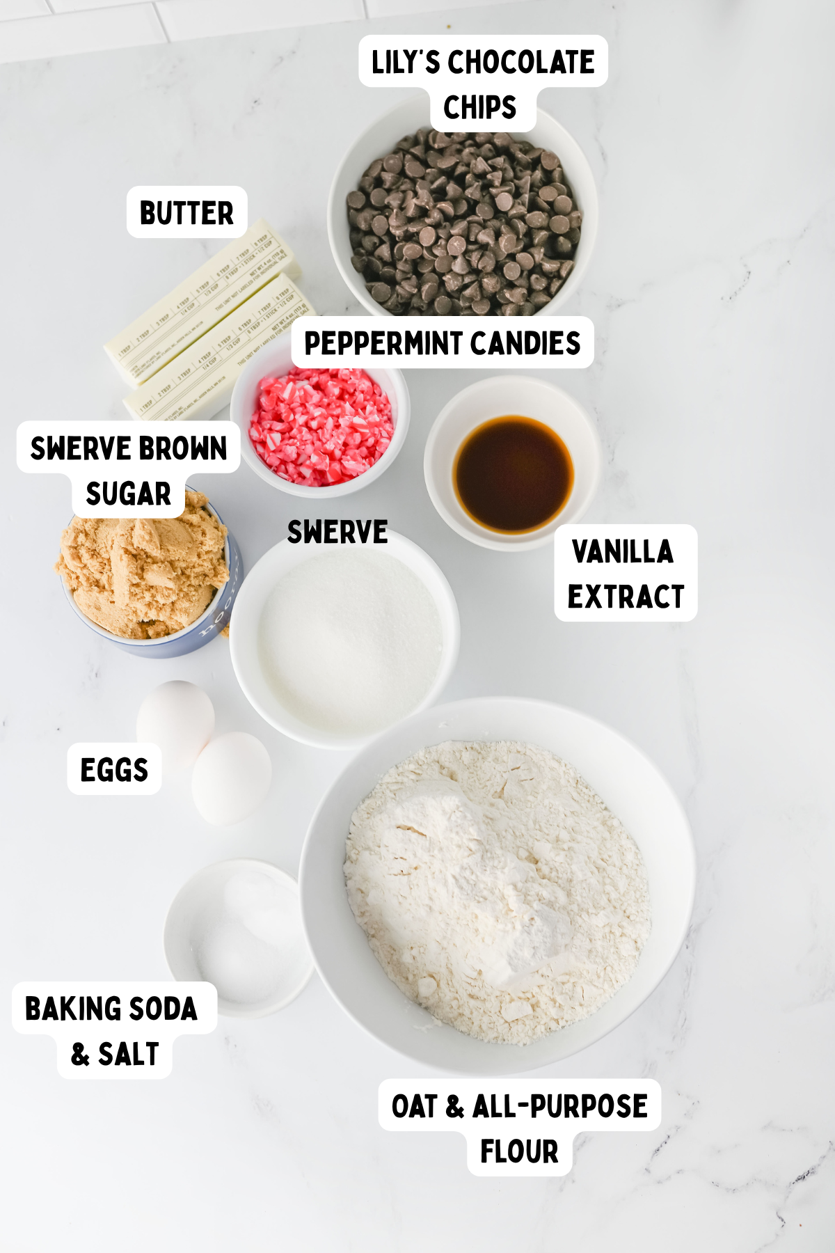 An overhead view of labeled ingredients in bowls and on a marble surface, including butter, eggs, swerve brown sugar, swerve, oat and all-purpose flour, baking soda and salt, vanilla extract, peppermint candies, and chocolate chips.