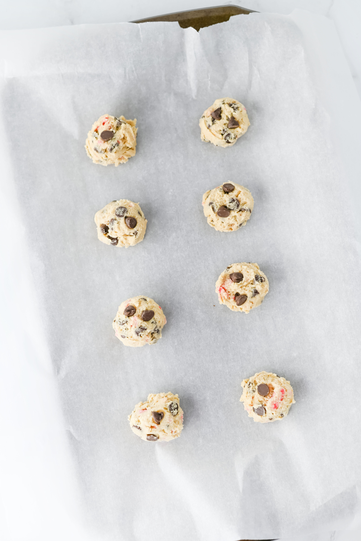 Eight scoops of chocolate chip cookie dough are arranged in two columns on a parchment-lined baking sheet, ready to be baked.