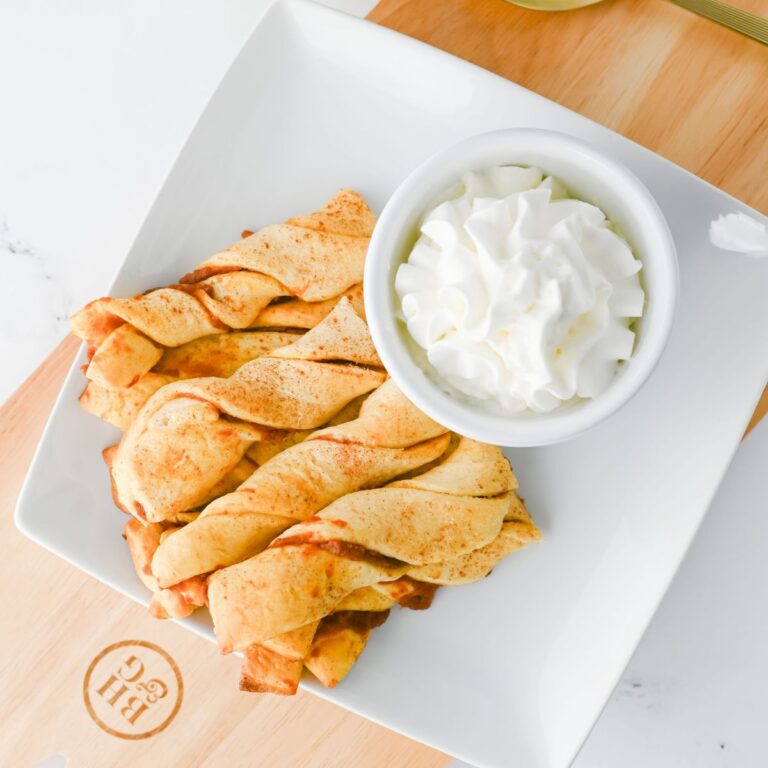 A white plate holds several twisted pastry sticks next to a small bowl of whipped cream. The plate is placed on a wooden cutting board with a logo on it.