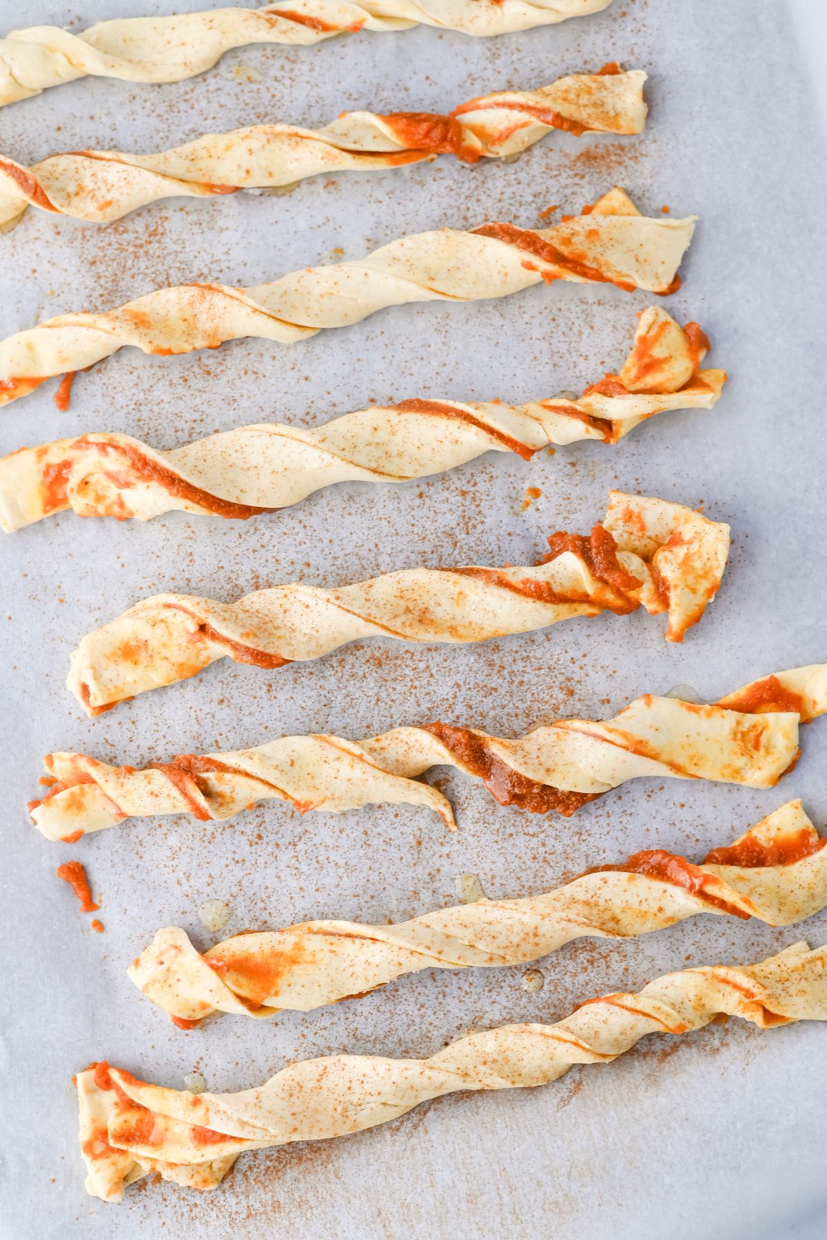 Several uncooked pastry twists with a light reddish sauce and sprinkled seasoning are arranged on a sheet of parchment paper. The pastries appear to be ready for baking.