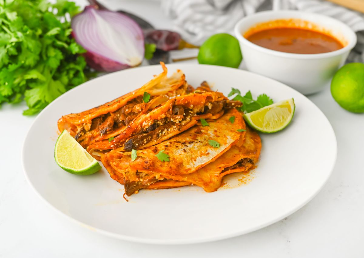 A white plate with folded orange-red birria tacos, garnished with lime wedges and cilantro, sits on a white surface. In the background are cilantro, a halved red onion, whole limes, and a bowl of reddish sauce.