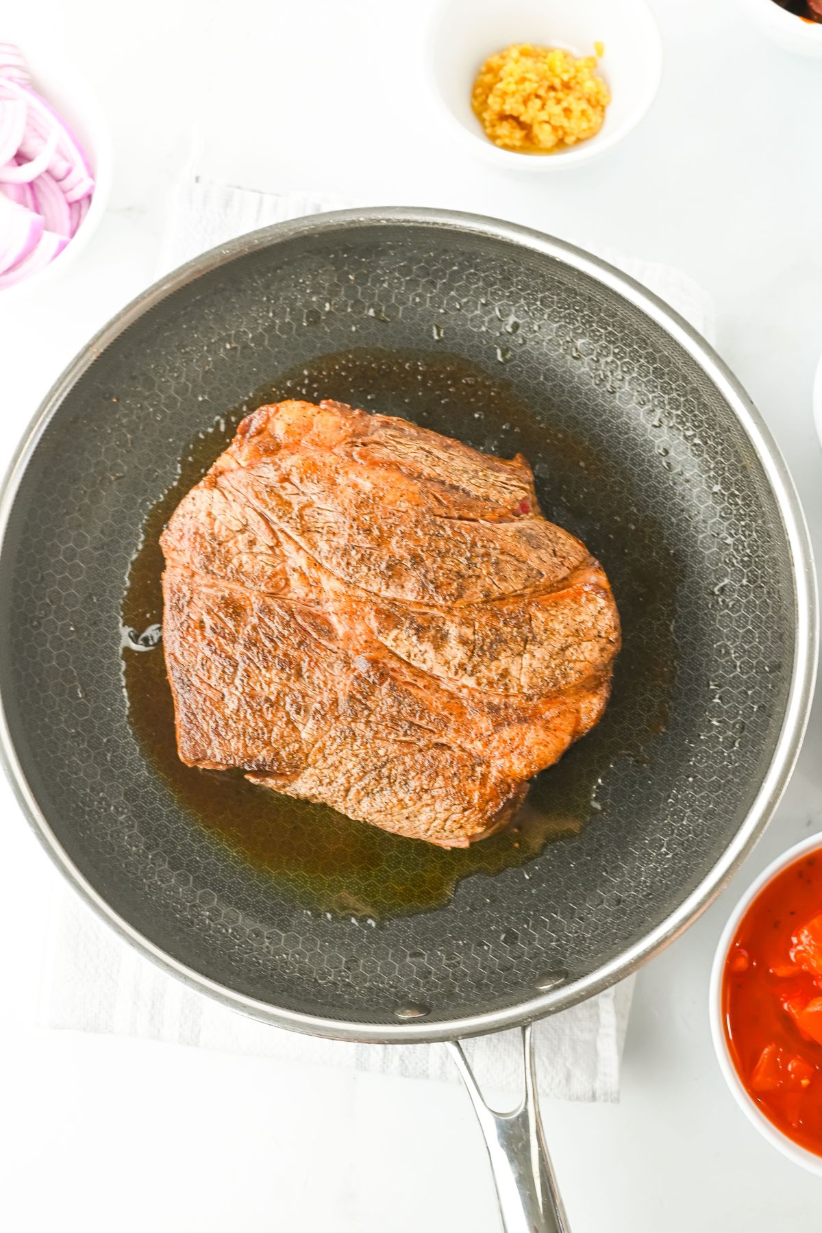 A seasoned piece of beef is searing in a black nonstick skillet with some oil. Surrounding the pan are small bowls containing sliced red onions, minced garlic, and red sauce.
