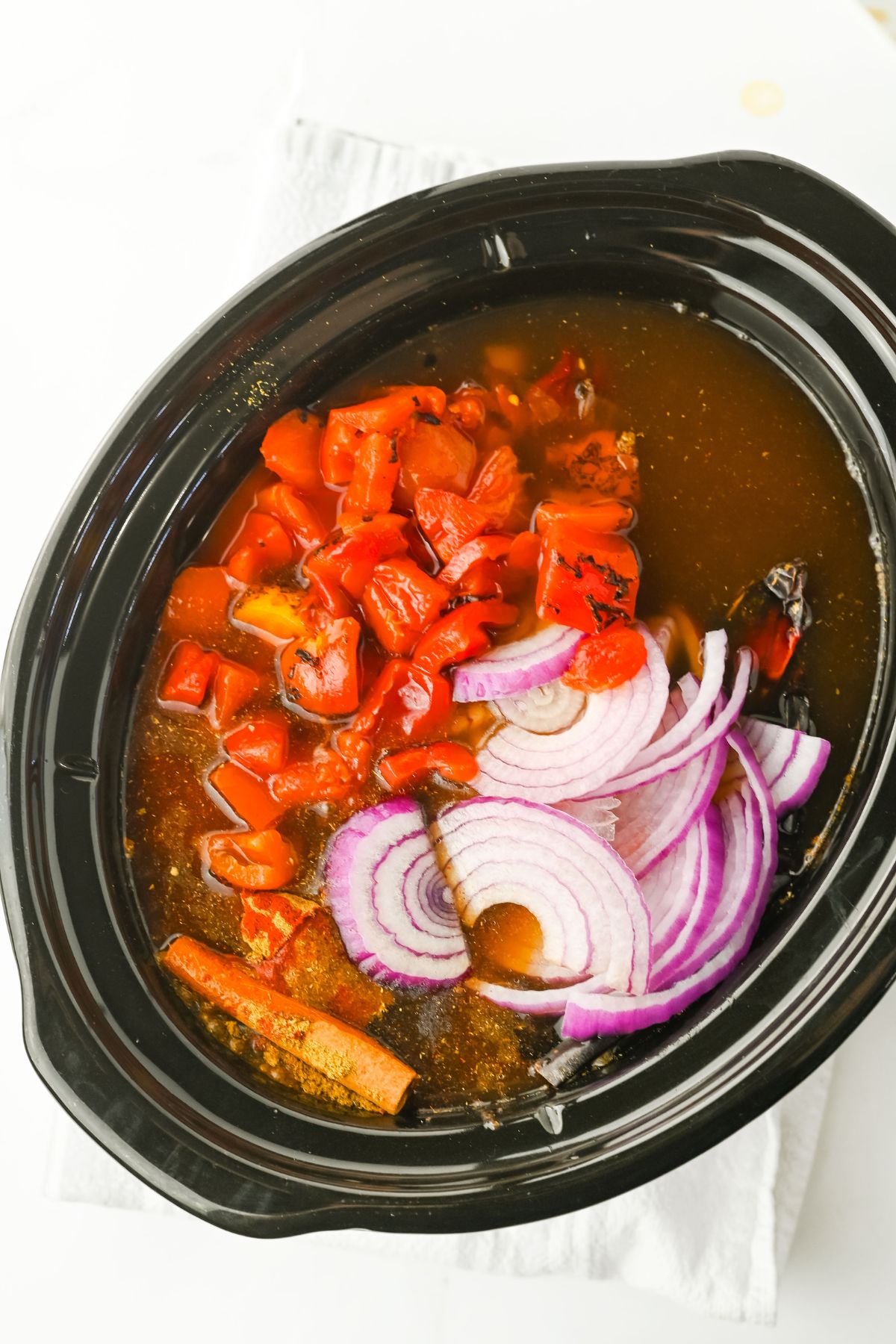 A black slow cooker filled with broth, chopped red bell peppers, sliced red onions, and a cinnamon stick, sitting on a white cloth.