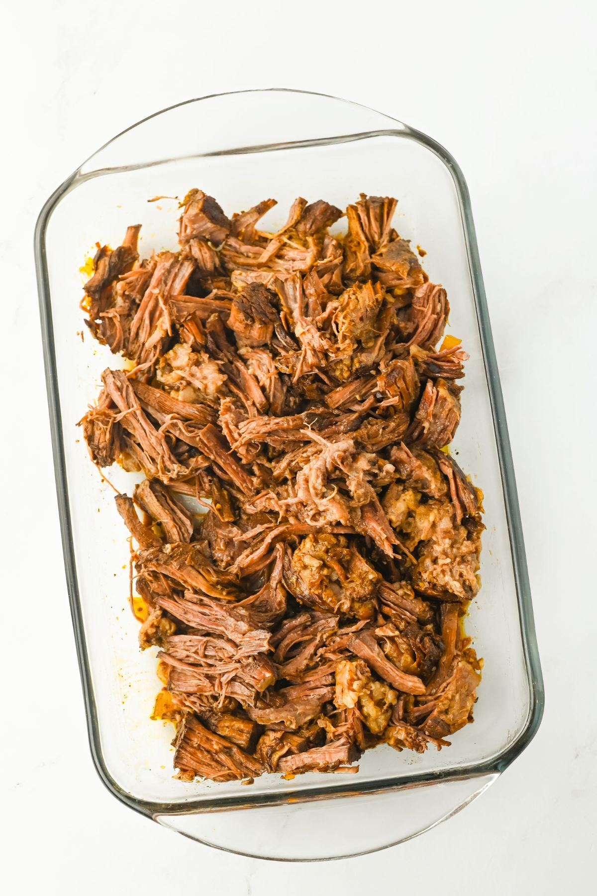 A glass baking dish filled with shredded cooked beef roast, arranged in an uneven layer on a white surface.