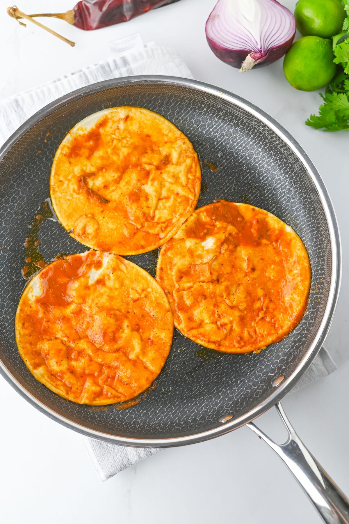 Three corn tortillas coated in red sauce are being heated in a nonstick skillet. A halved red onion, two limes, fresh cilantro, and a dried chili are visible in the background on a white surface.