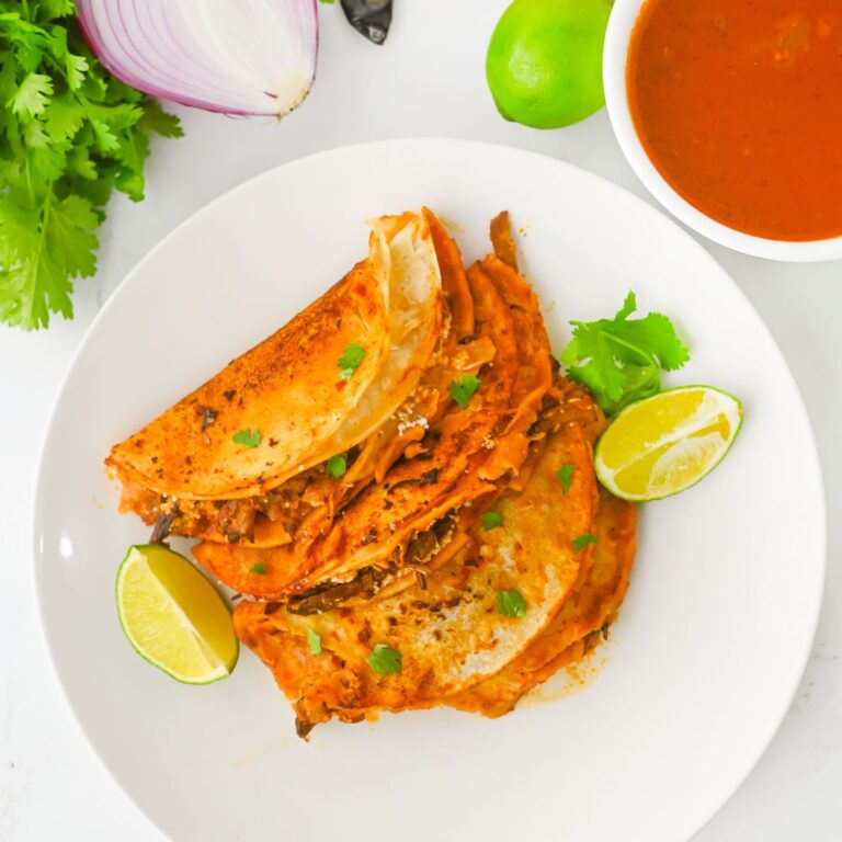 Three folded birria tacos with cilantro and lime wedges are served on a white plate. A bowl of red sauce, half a red onion, cilantro, and a whole lime are visible nearby on a white surface.