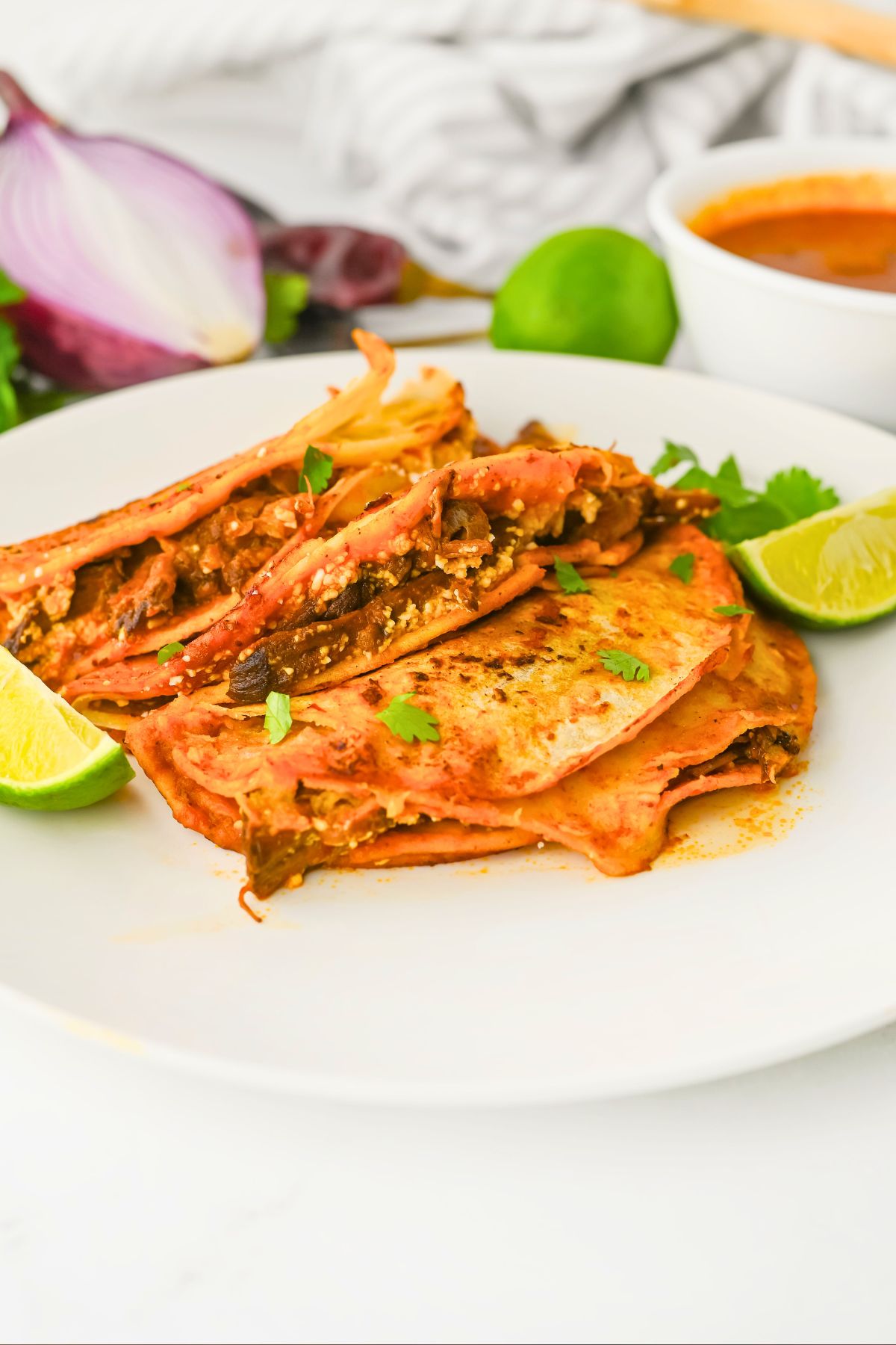 A white plate holds three folded, saucy birria tacos garnished with chopped cilantro and lime wedges. A sliced red onion, whole lime, and a bowl of broth are in the background.
