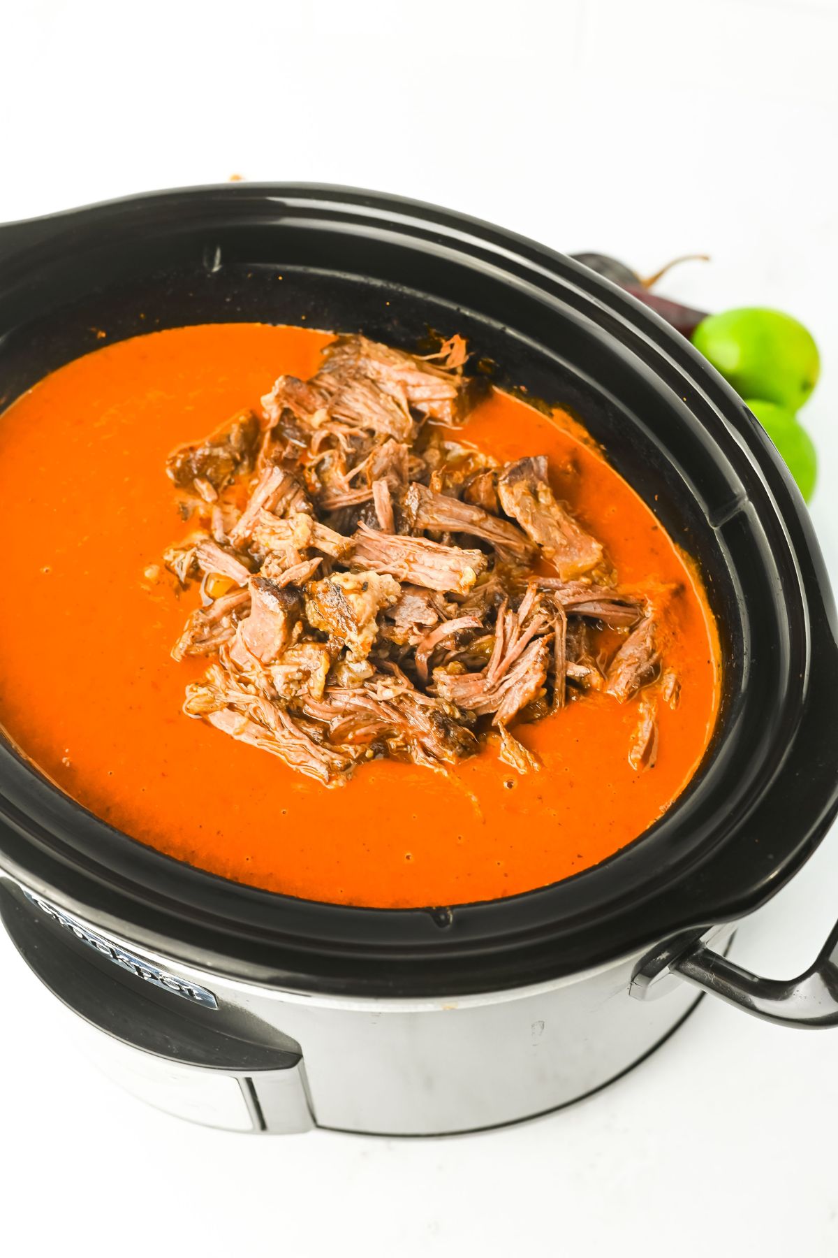 A black slow cooker filled with shredded beef in a red sauce. The slow cooker is on a white surface with two whole limes placed in the background.