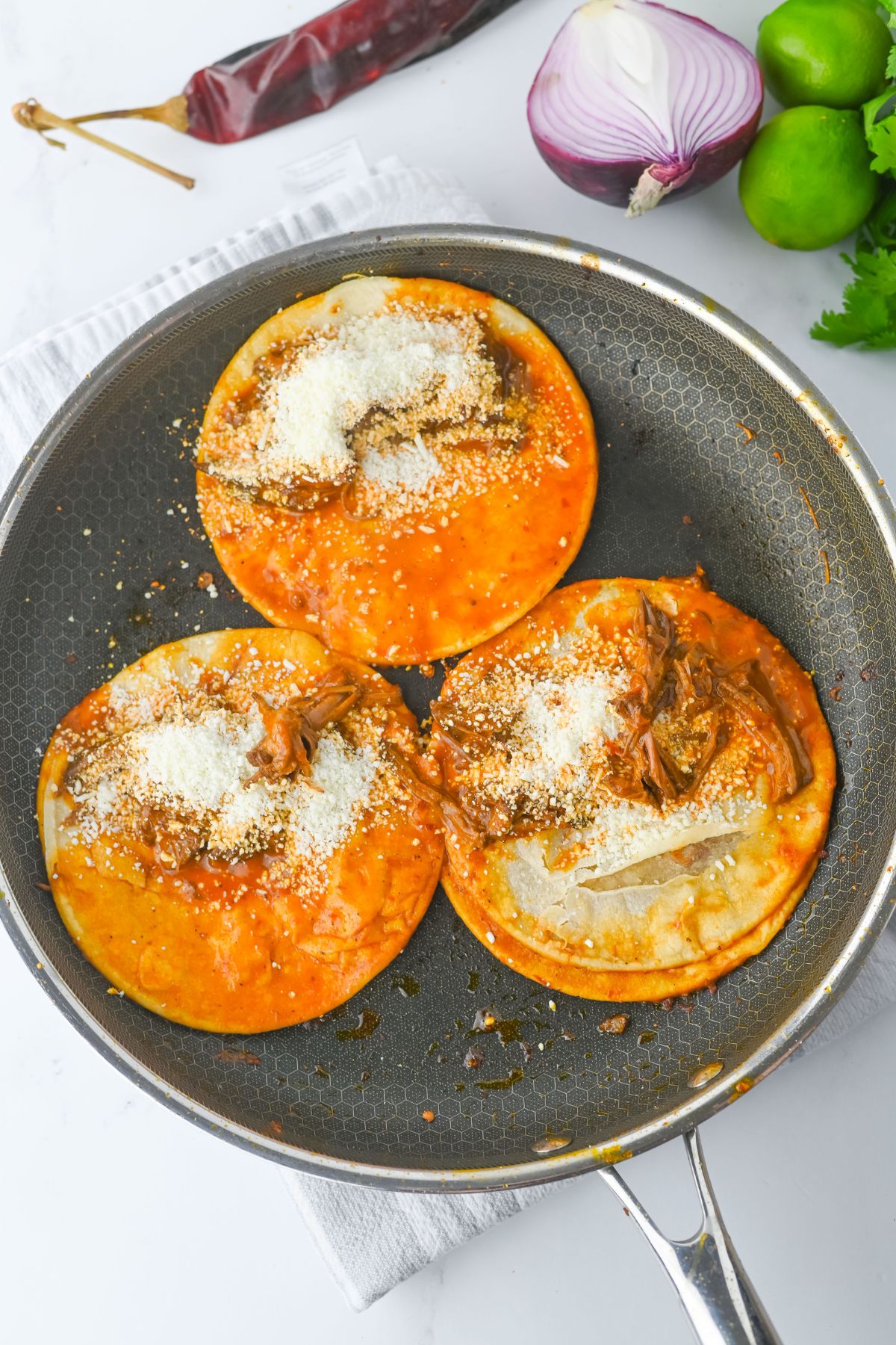 Three corn tortillas coated in red sauce and topped with shredded meat and grated cheese are in a frying pan. A half red onion, whole limes, cilantro, and a dried chili are visible in the background.